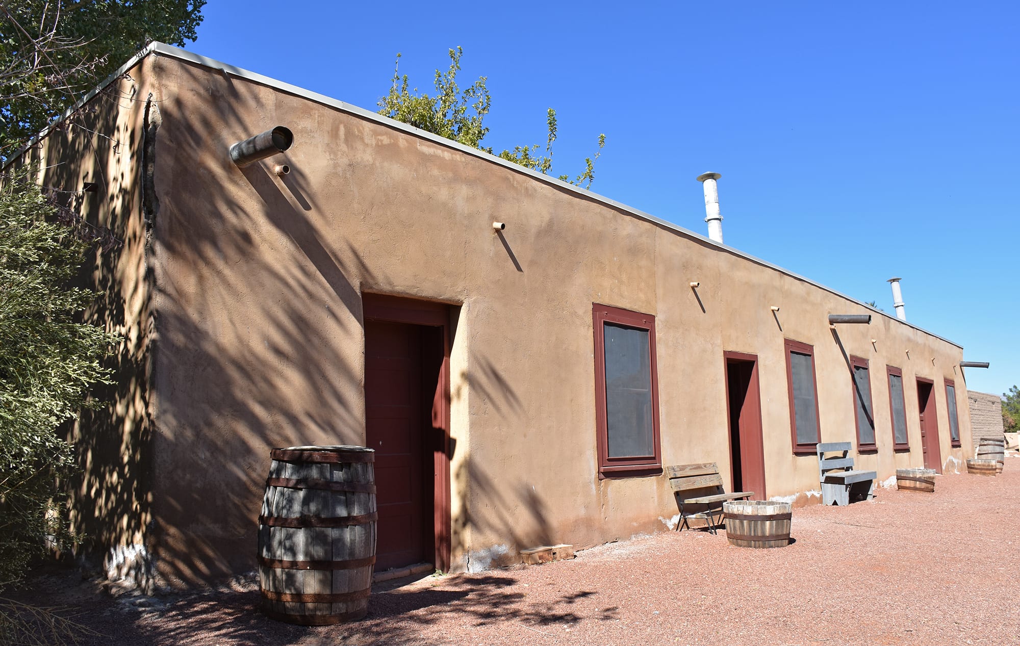 A long single-storey adobe building with red-brown doors and windows, wooden barrels and benches outside, under a clear blue sky