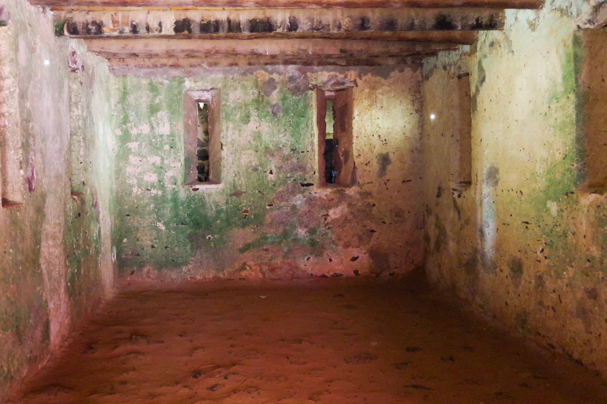 A bare, low room with stained green and pink walls, a beamed ceiling and two narrow slit windows