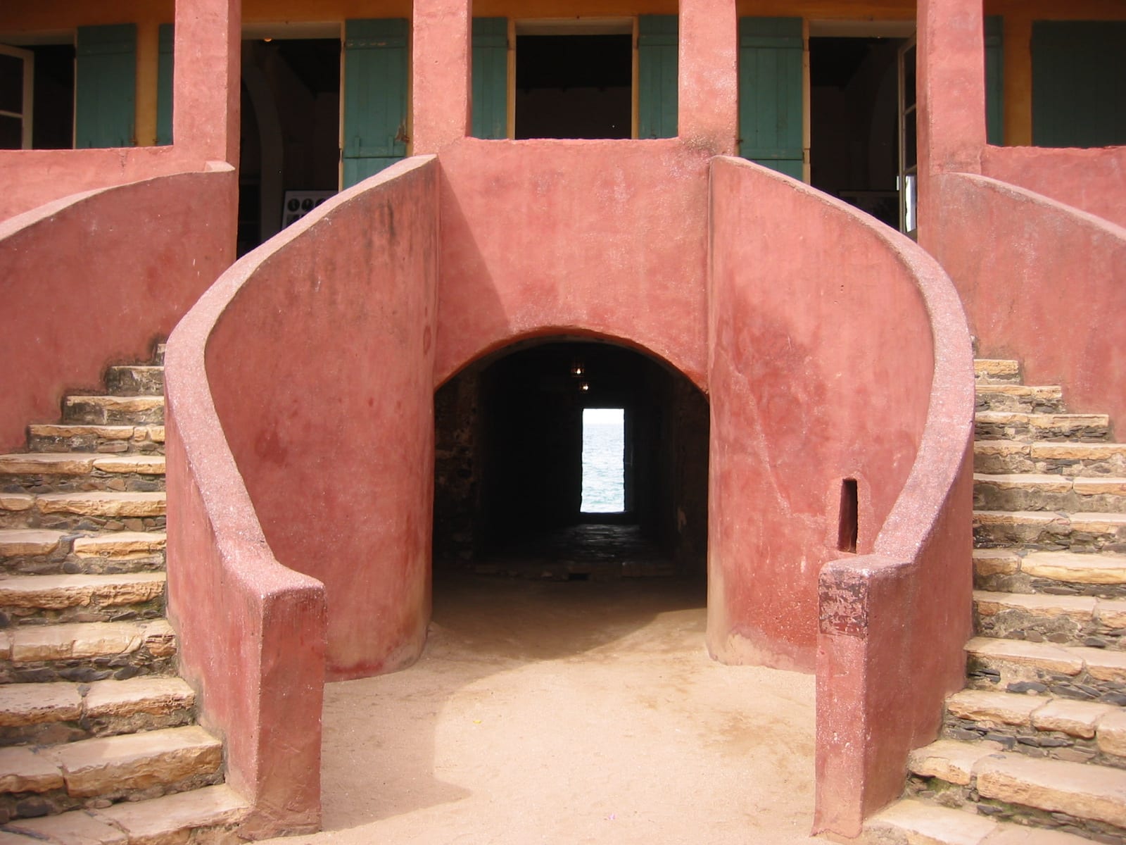 Two curved pink staircases framing an arched passage that leads through a dark corridor to a small bright doorway open to the sea