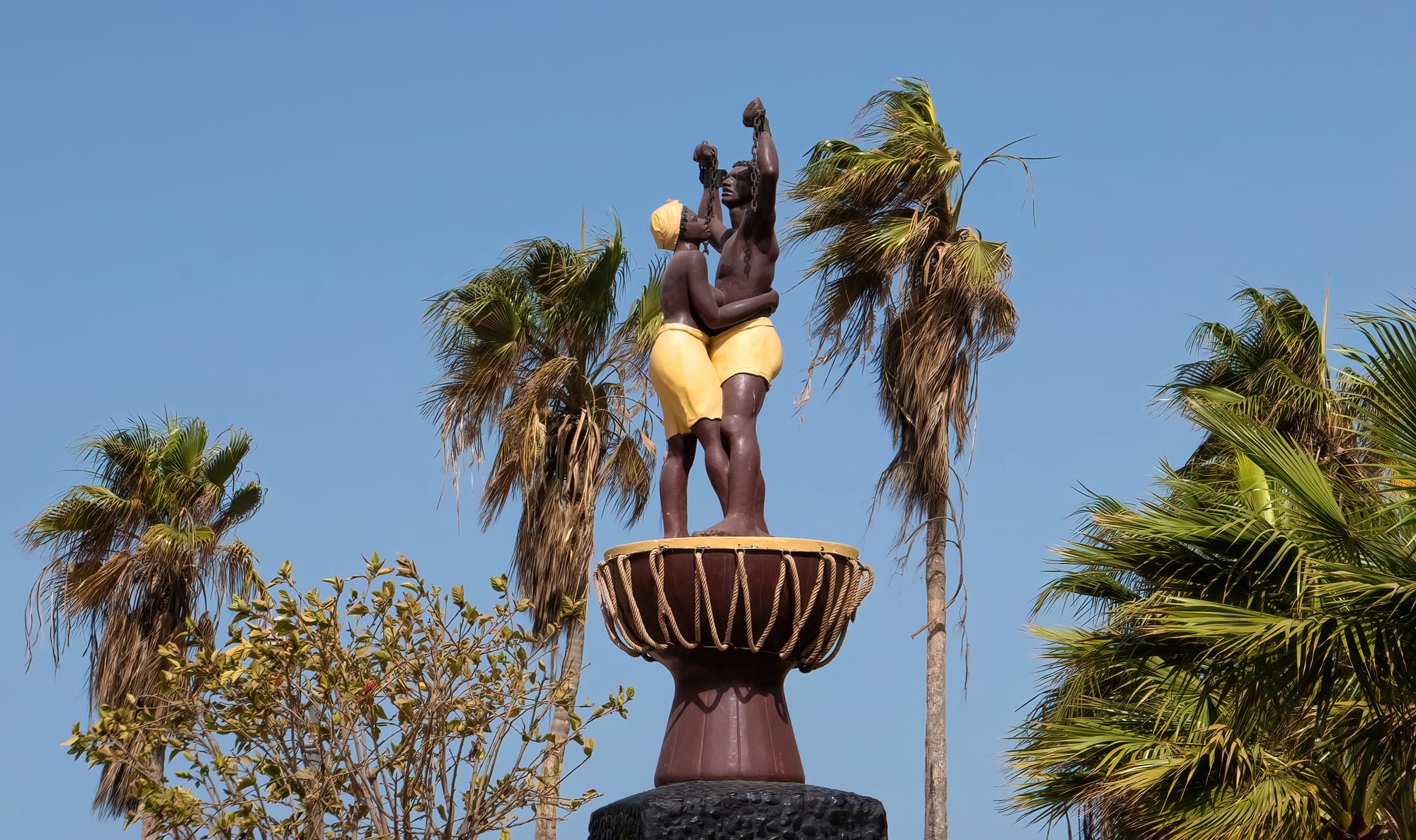 A painted statue of a man raising his arms with broken chains, embracing a woman, on a tall drum-shaped pedestal among palm trees