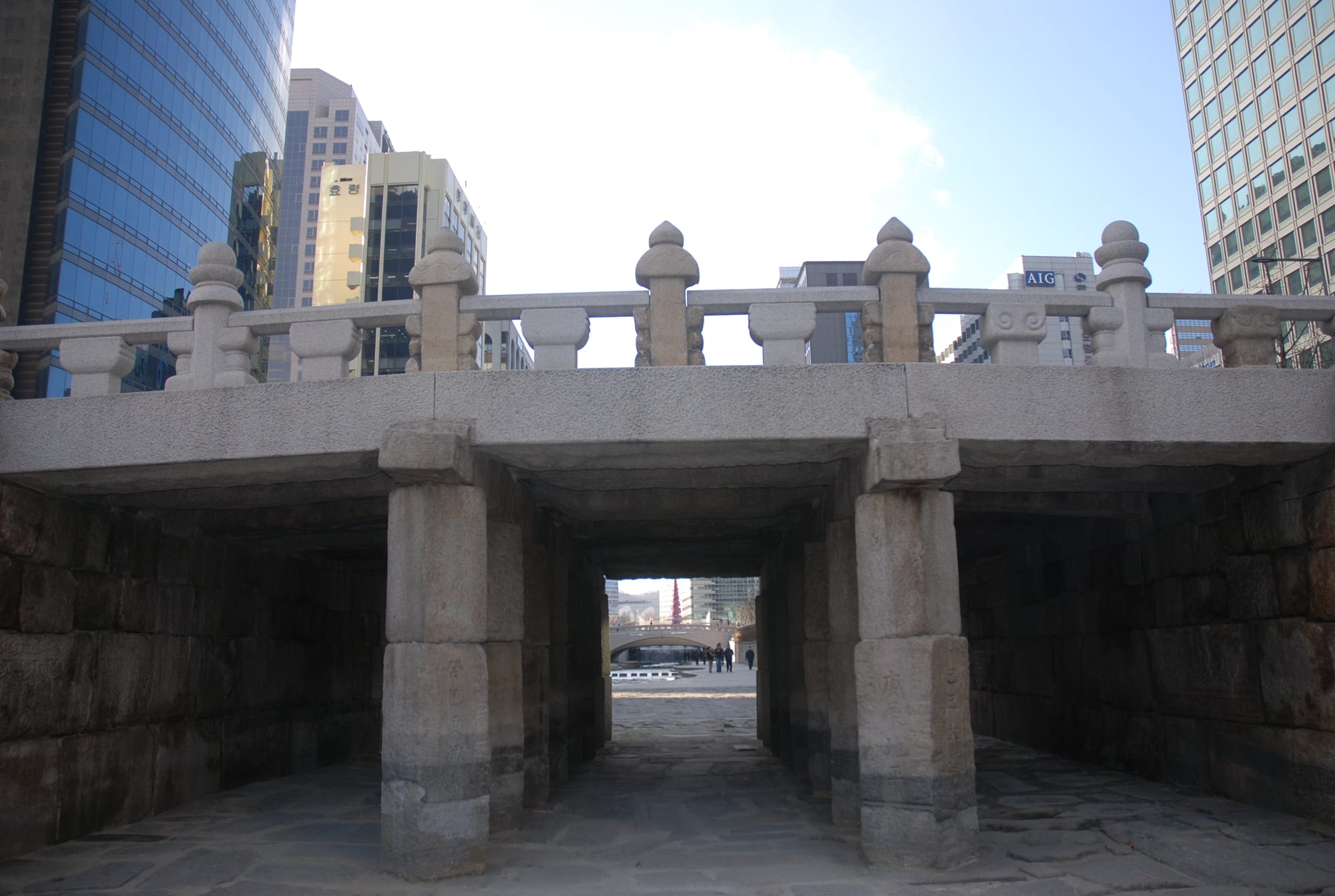 A low stone bridge with carved stone railings and square stone piers spanning a paved stream bed, with glass office towers behind