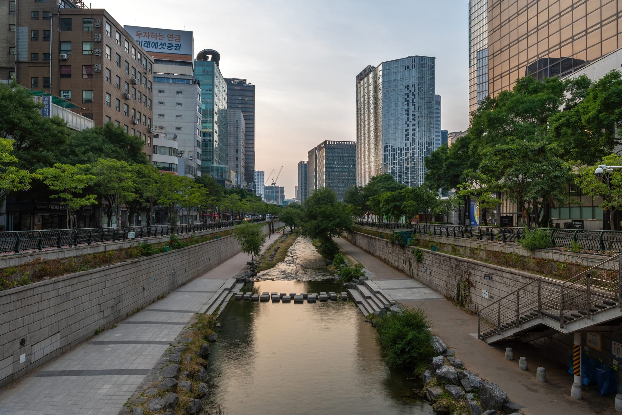 A shallow stream with stepping stones running between stone embankments and footpaths, lined with trees and overlooked by office towers