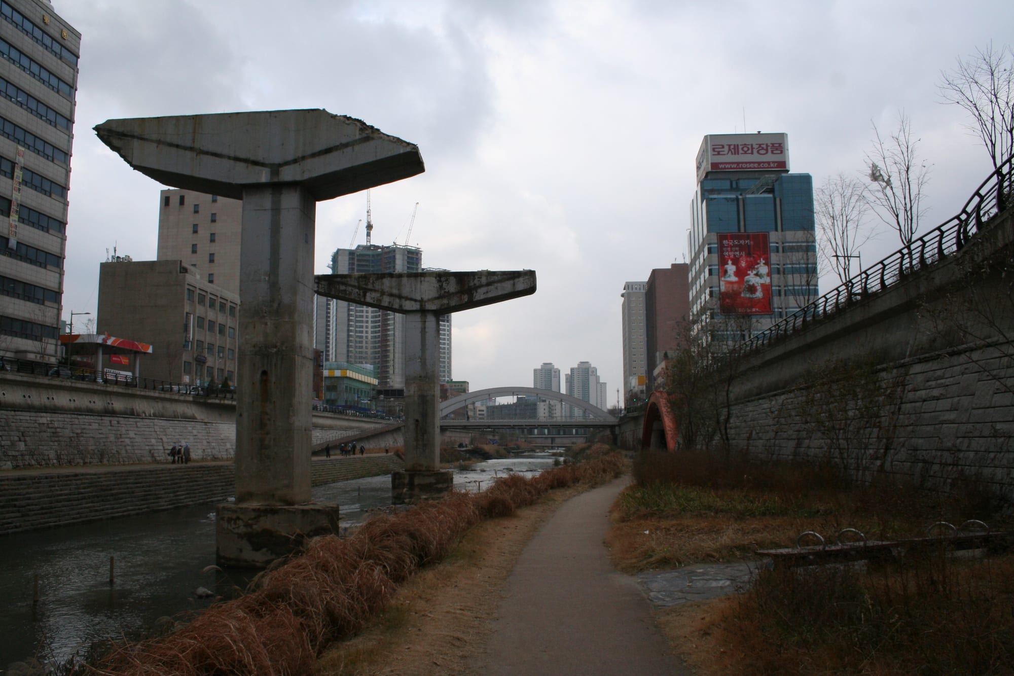 Two tall concrete pillars with flared tops standing beside a stream and a footpath, with apartment towers and an arched bridge in the distance