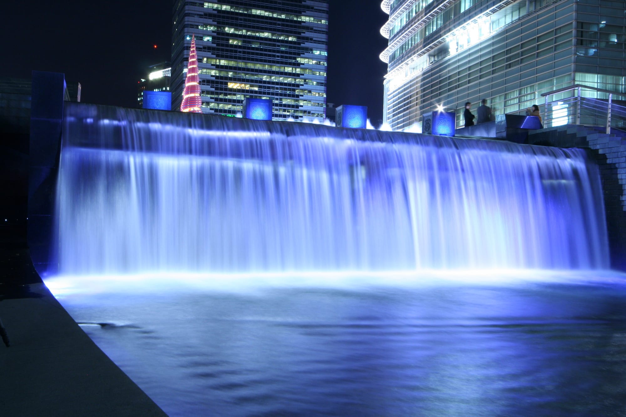 A wide curtain of water lit blue at night pouring into a pool, with a red spiral sculpture and lit office towers behind