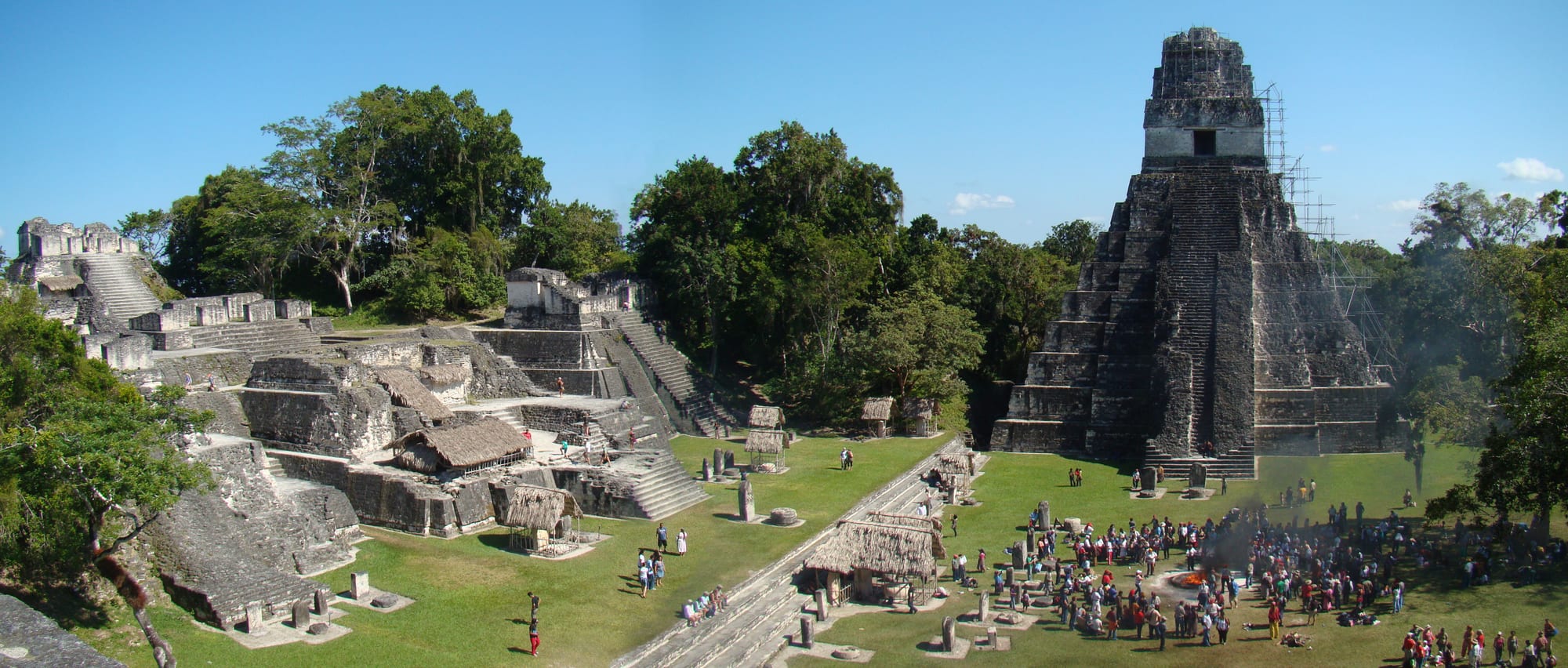 A wide grassy plaza with a crowd gathered near a smoking fire, a tall steep stone pyramid with a roof comb on the right and terraced stone buildings on the left