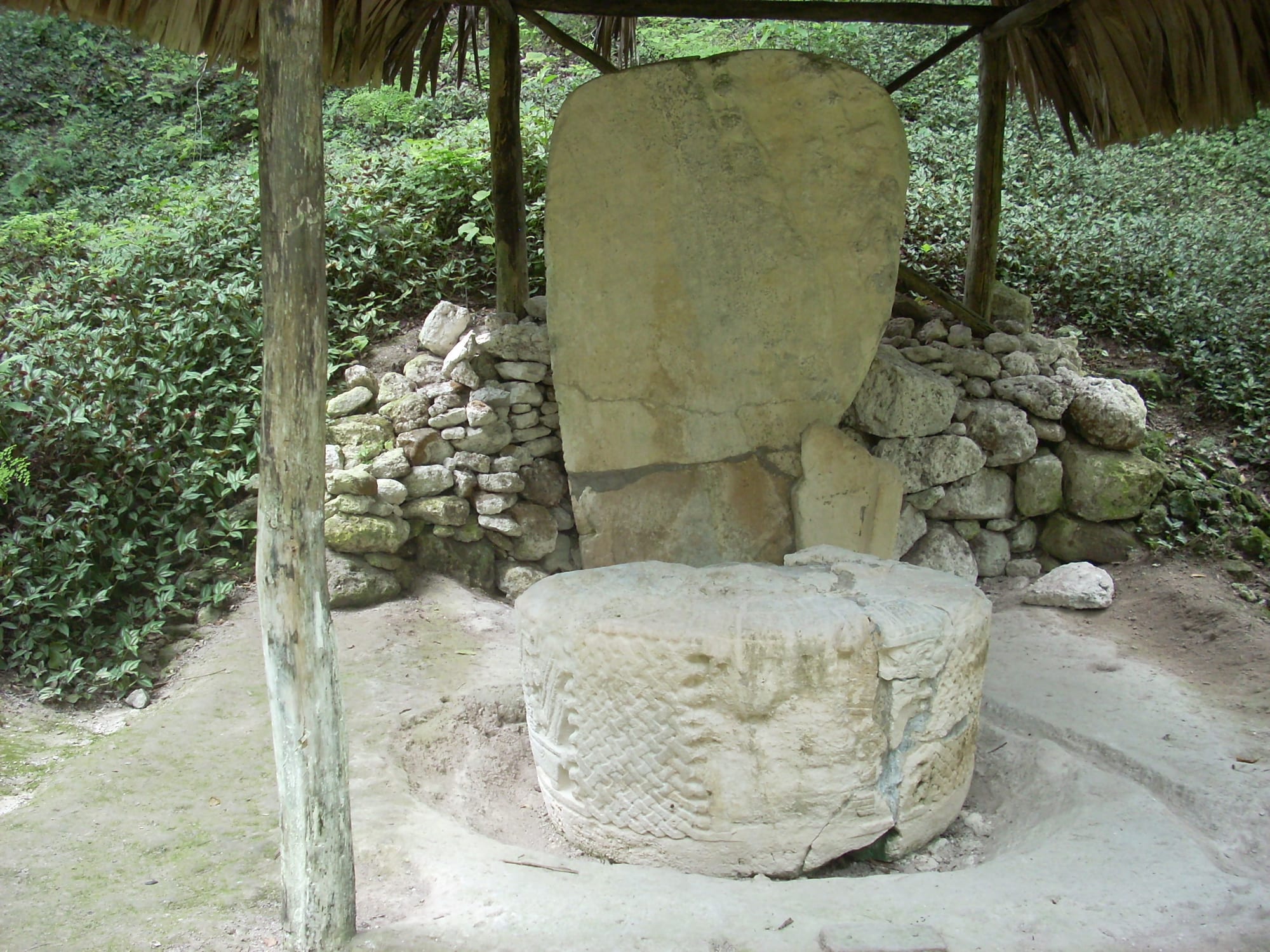 An upright weathered stone slab with a round carved stone altar in front of it, under a thatched shelter in the forest