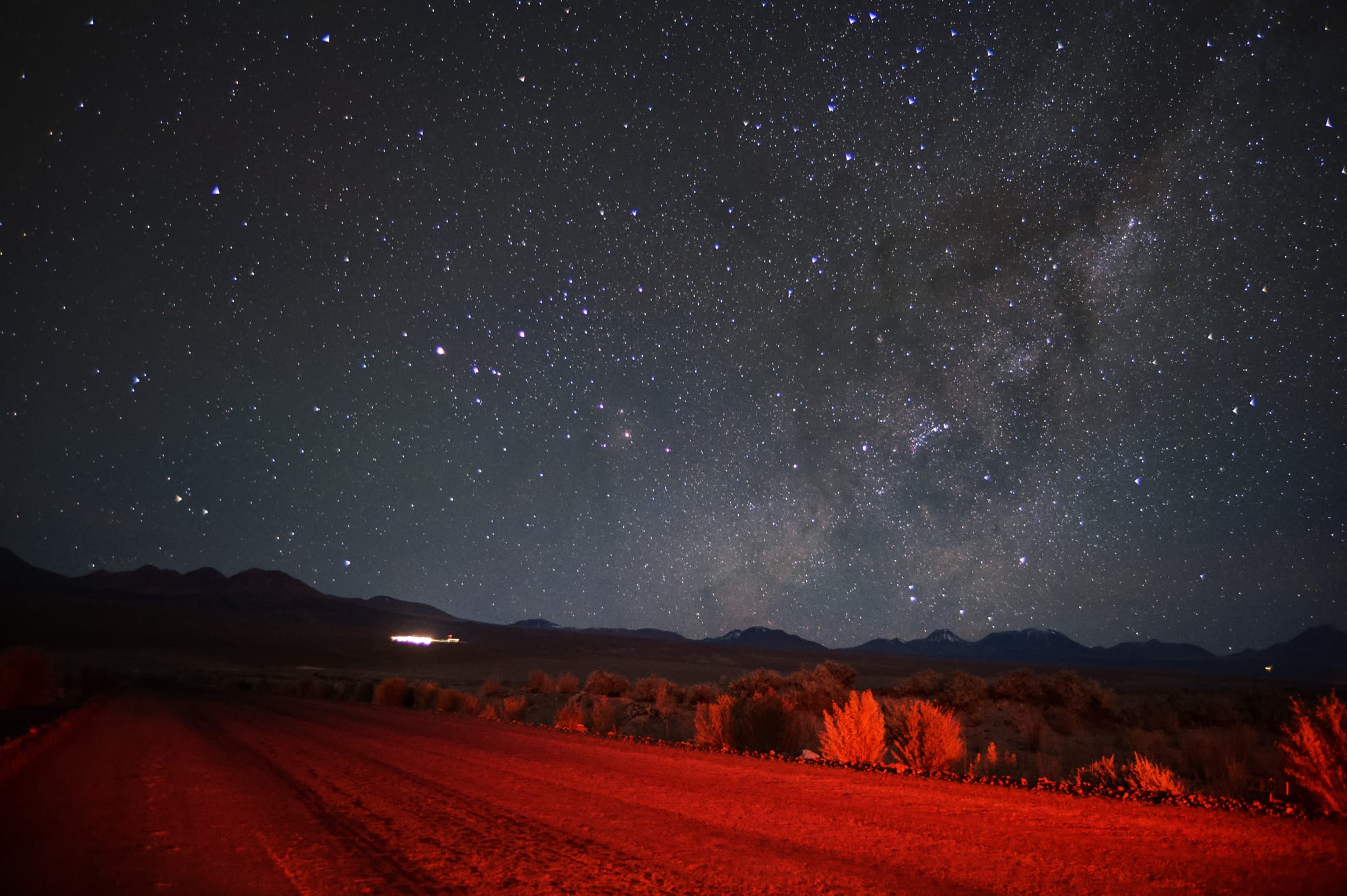 A night sky crowded with stars and the faint band of the Milky Way above dark mountains, with a desert road and shrubs lit red in the foreground