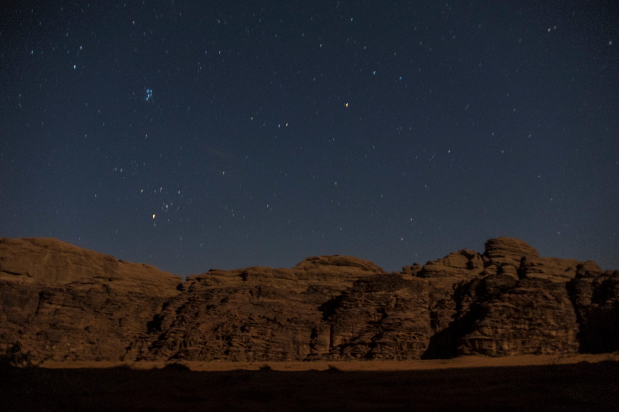 Weathered sandstone cliffs lit faintly against a deep blue night sky scattered with stars, above a dark sandy valley floor