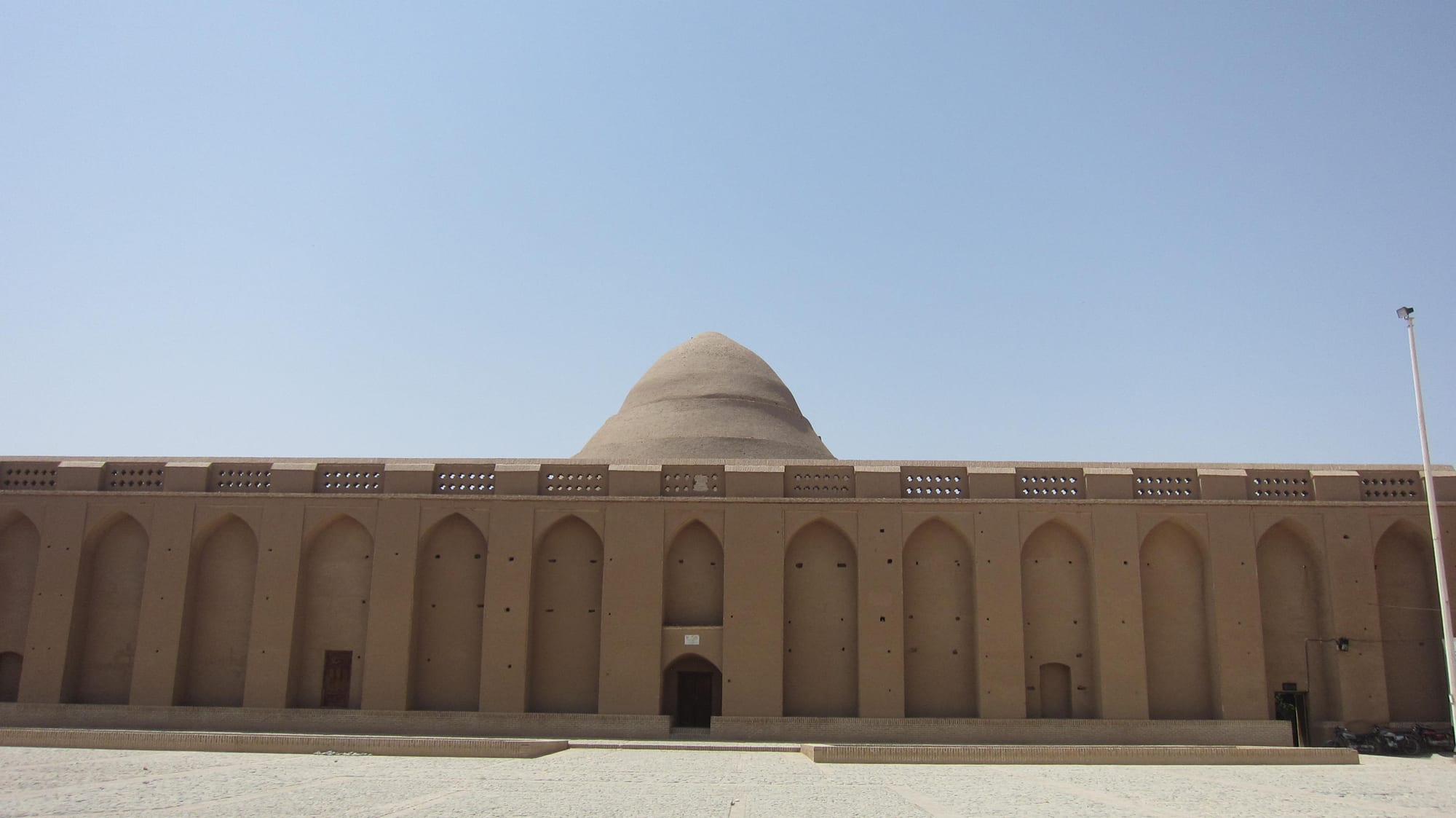 A tall stepped mud-brick dome rising behind a long wall of arched recesses under a pale blue sky