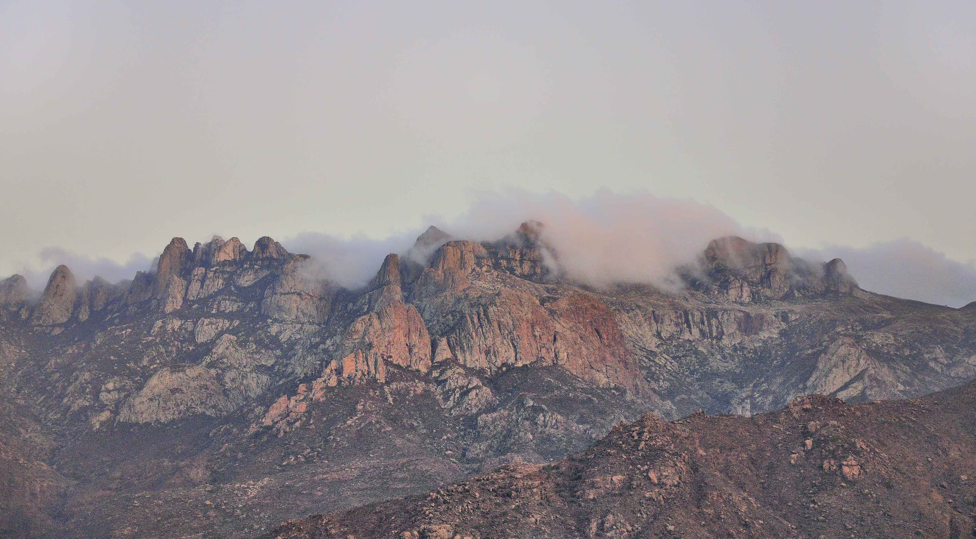 Jagged pink and grey granite peaks partly wrapped in low cloud under a pale sky