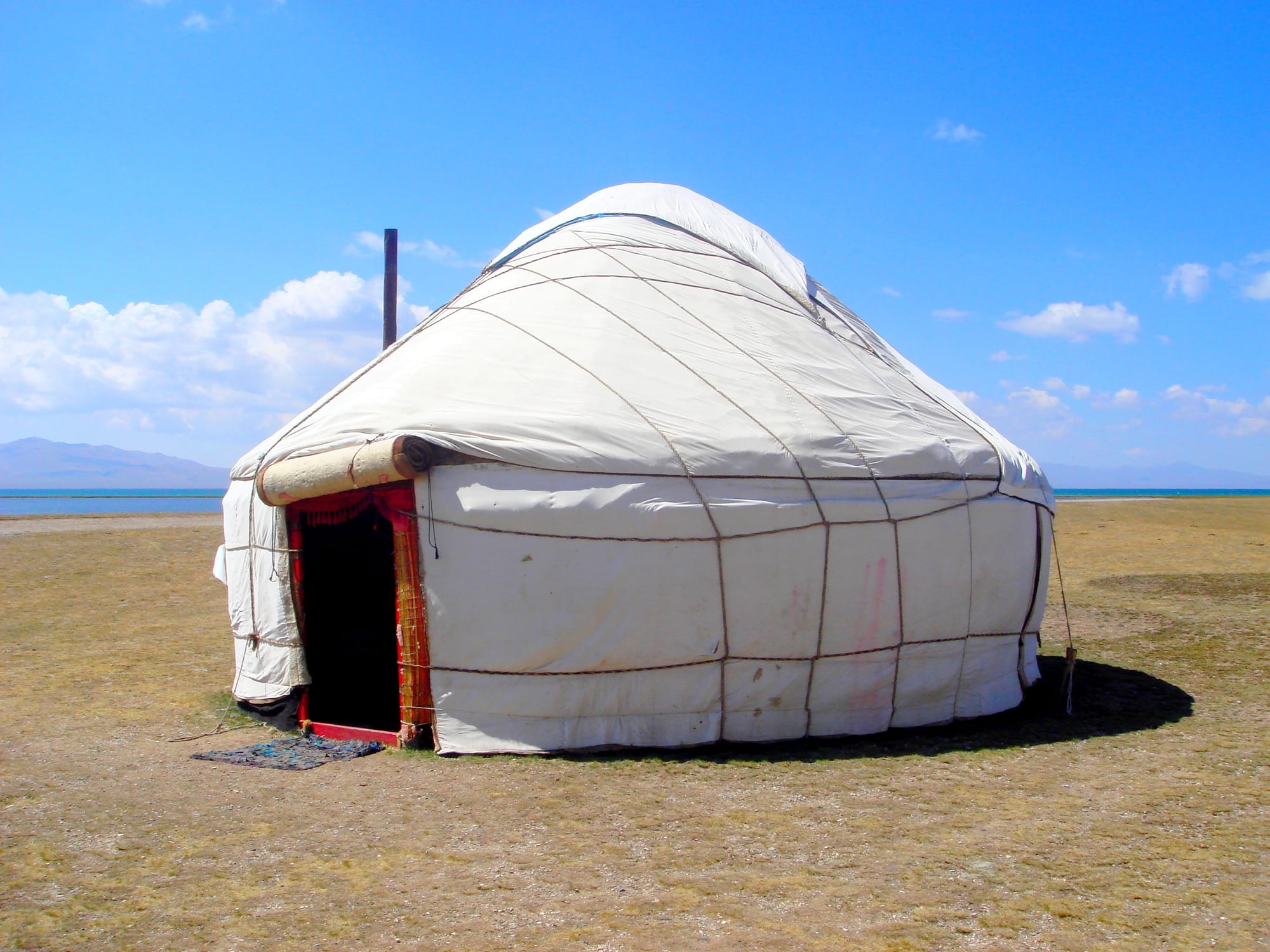 A round white felt tent with a red-framed doorway standing on dry grassland beside a blue lake, with mountains on the horizon