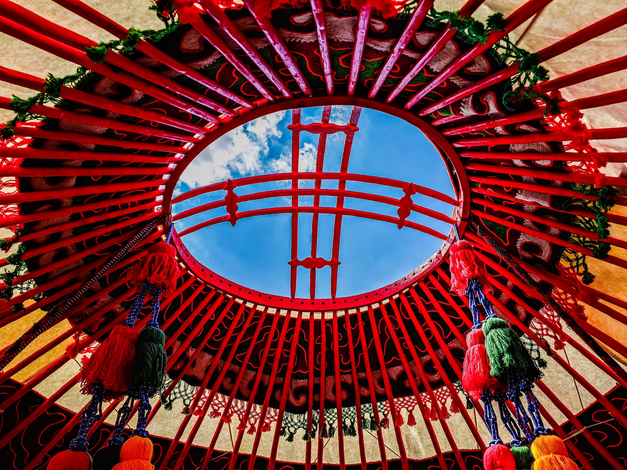 Looking straight up inside a yurt at a red wooden crown ring, with red roof poles radiating from it and blue sky visible through the opening