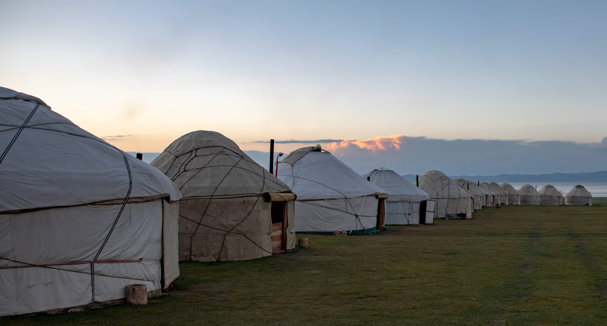A long row of white and cream felt yurts on green grass at dusk, with a lake and distant hills behind