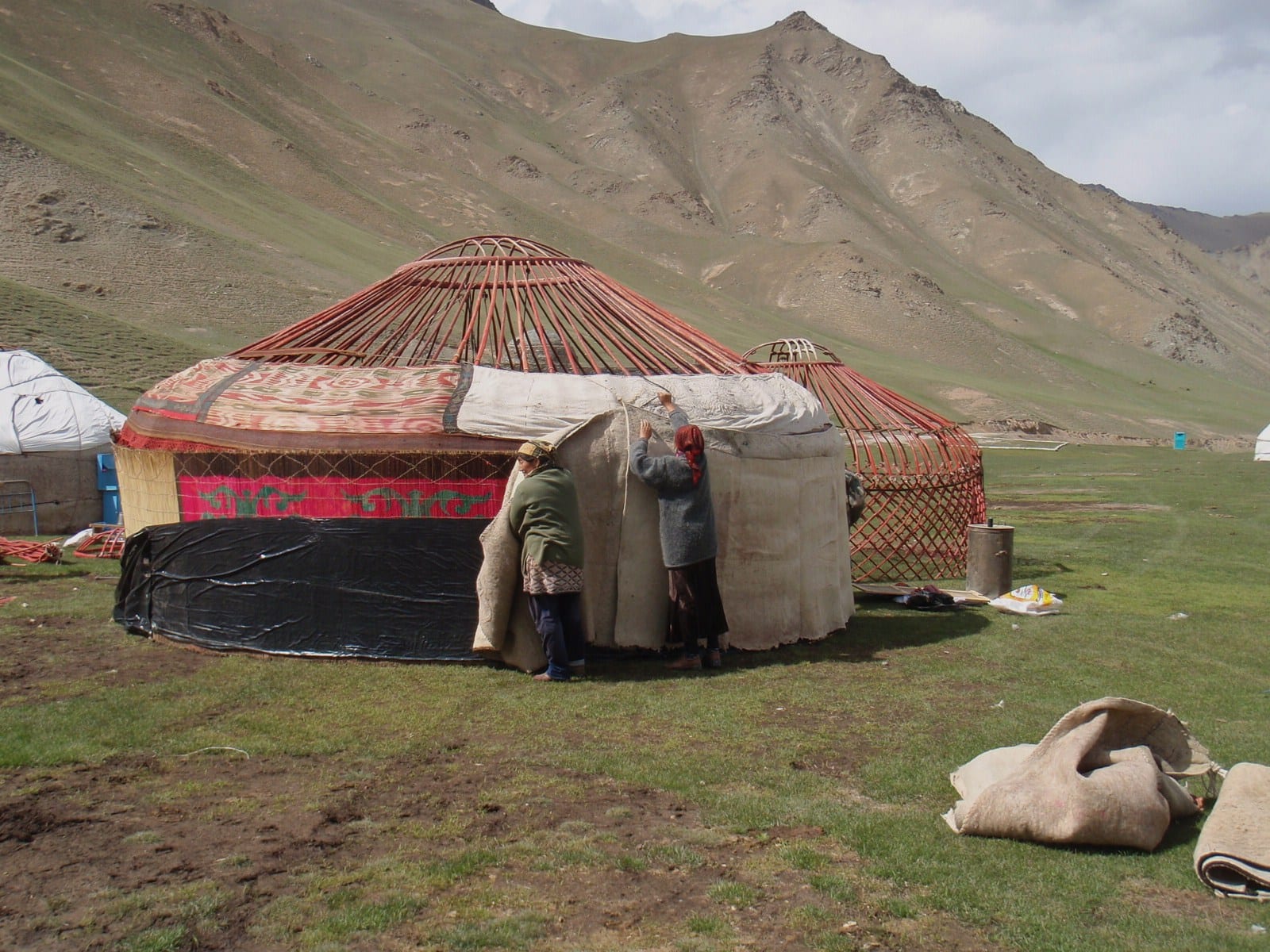 Two women pulling a large grey felt sheet over the red wooden frame of a yurt on a grassy valley floor, with a bare mountain behind