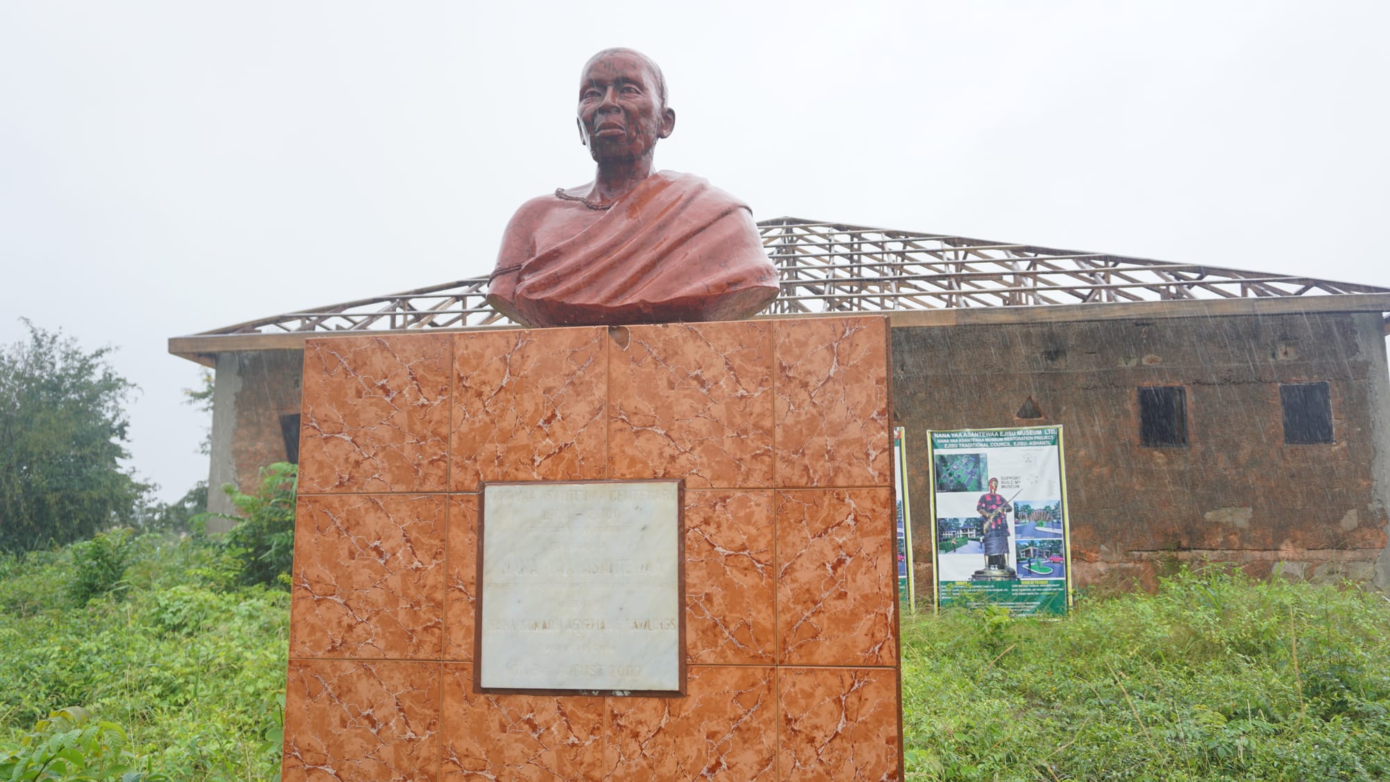 A reddish bust of an elderly woman wrapped in cloth, on a tiled pedestal with a plaque, in front of a building under construction