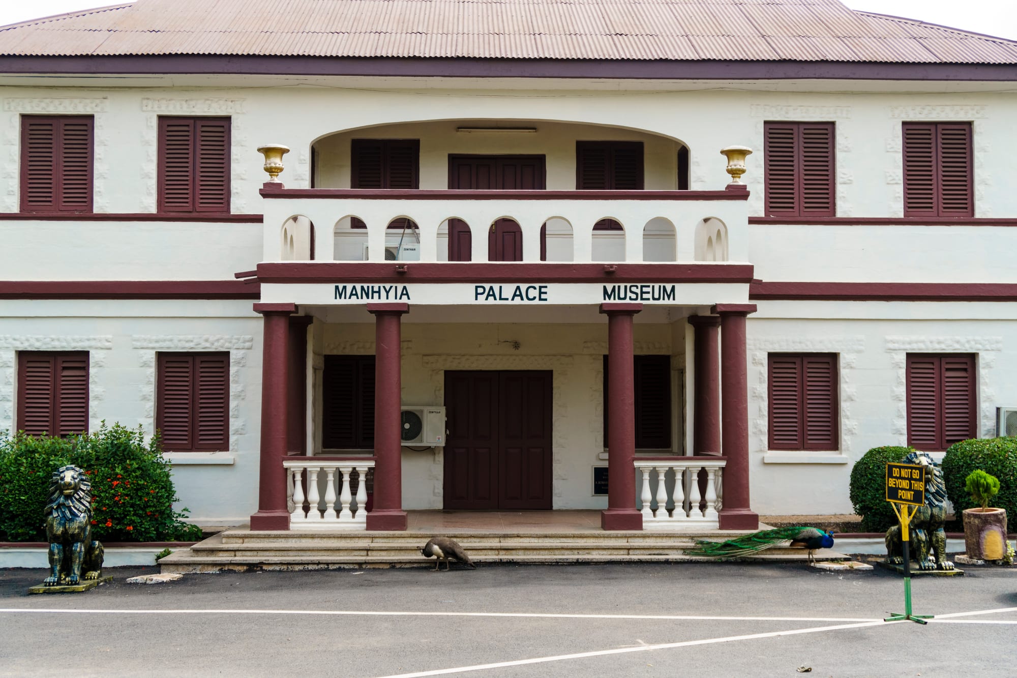 A white two-storey colonial building with maroon shutters, a columned porch and a sign reading Manhyia Palace Museum, with lion statues and peacocks in front