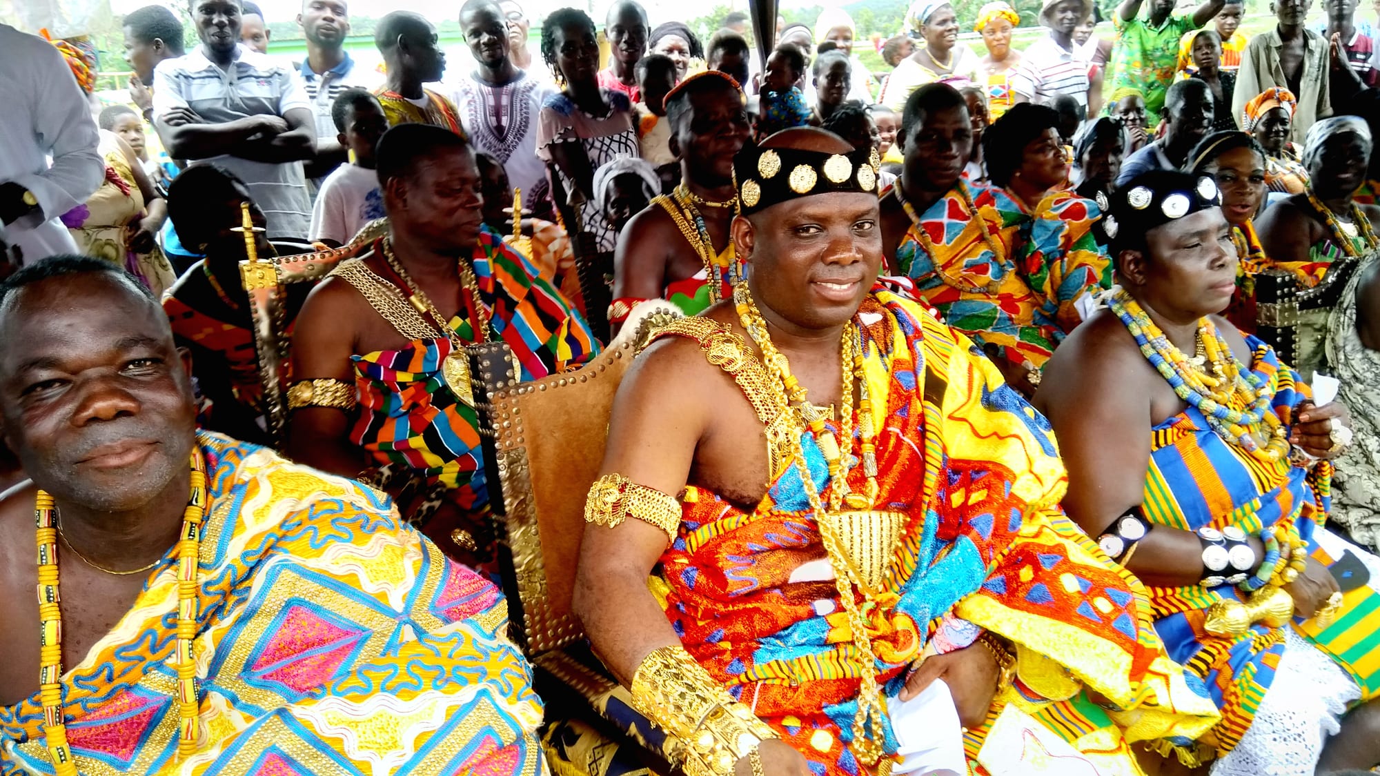 Seated Asante chiefs and elders in brightly patterned kente cloth with heavy gold necklaces, armlets and headbands, with a crowd standing behind them