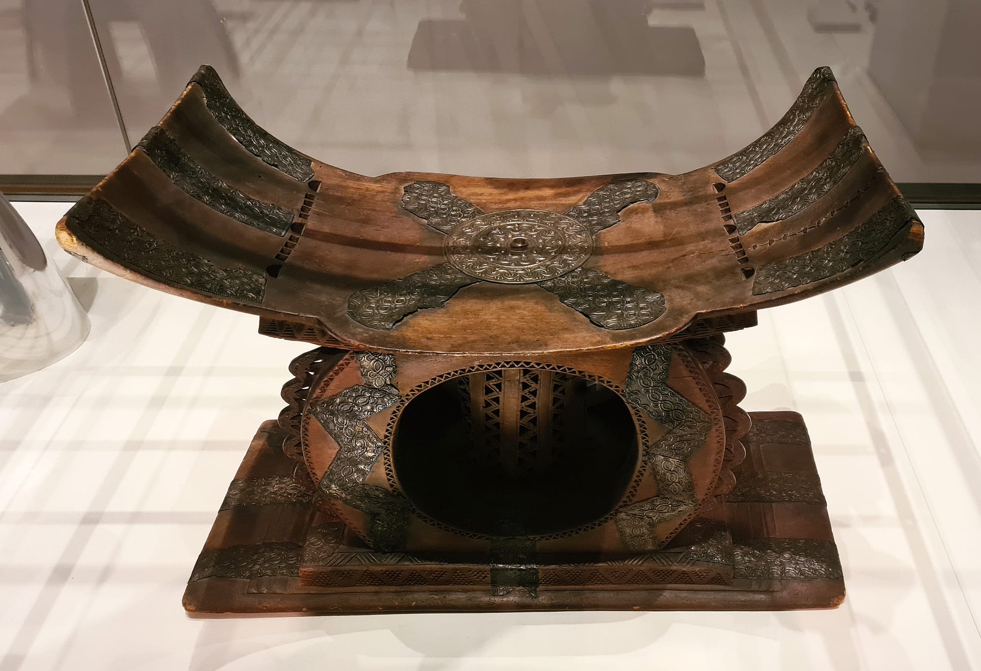 A carved wooden stool with a deeply curved seat, a round pierced central support and a flat rectangular base, decorated with patterned sheets of silver