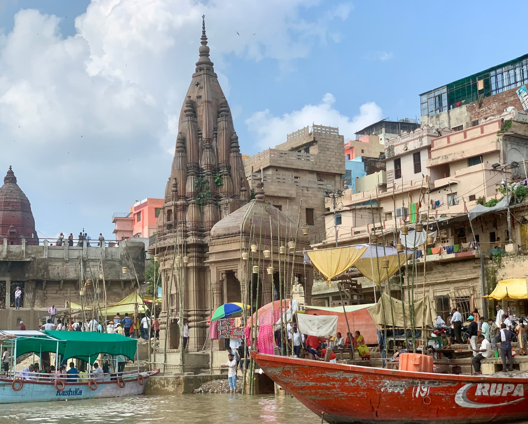 A tall carved stone temple spire rising beside steps crowded with people, boats and stacked bamboo poles on the riverbank