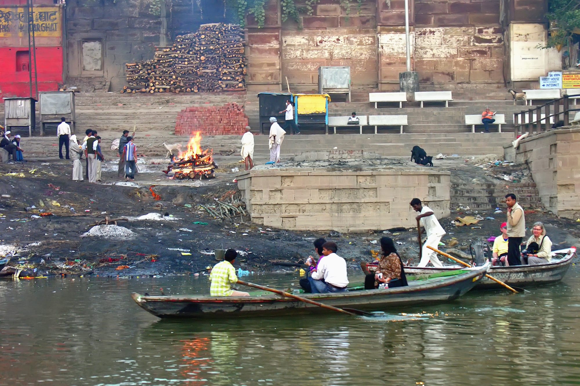 A funeral pyre burning on a riverbank below stone steps and stacks of firewood, with rowing boats passing in the foreground