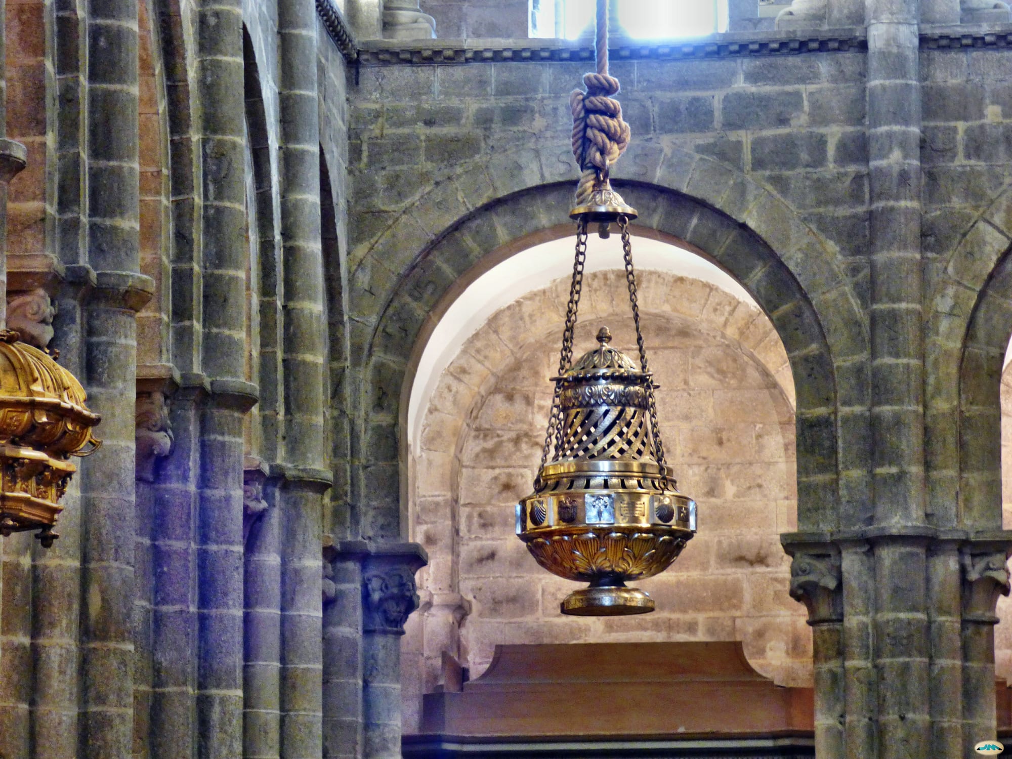 A large ornate brass incense burner hanging on chains from a thick rope between Romanesque stone arches