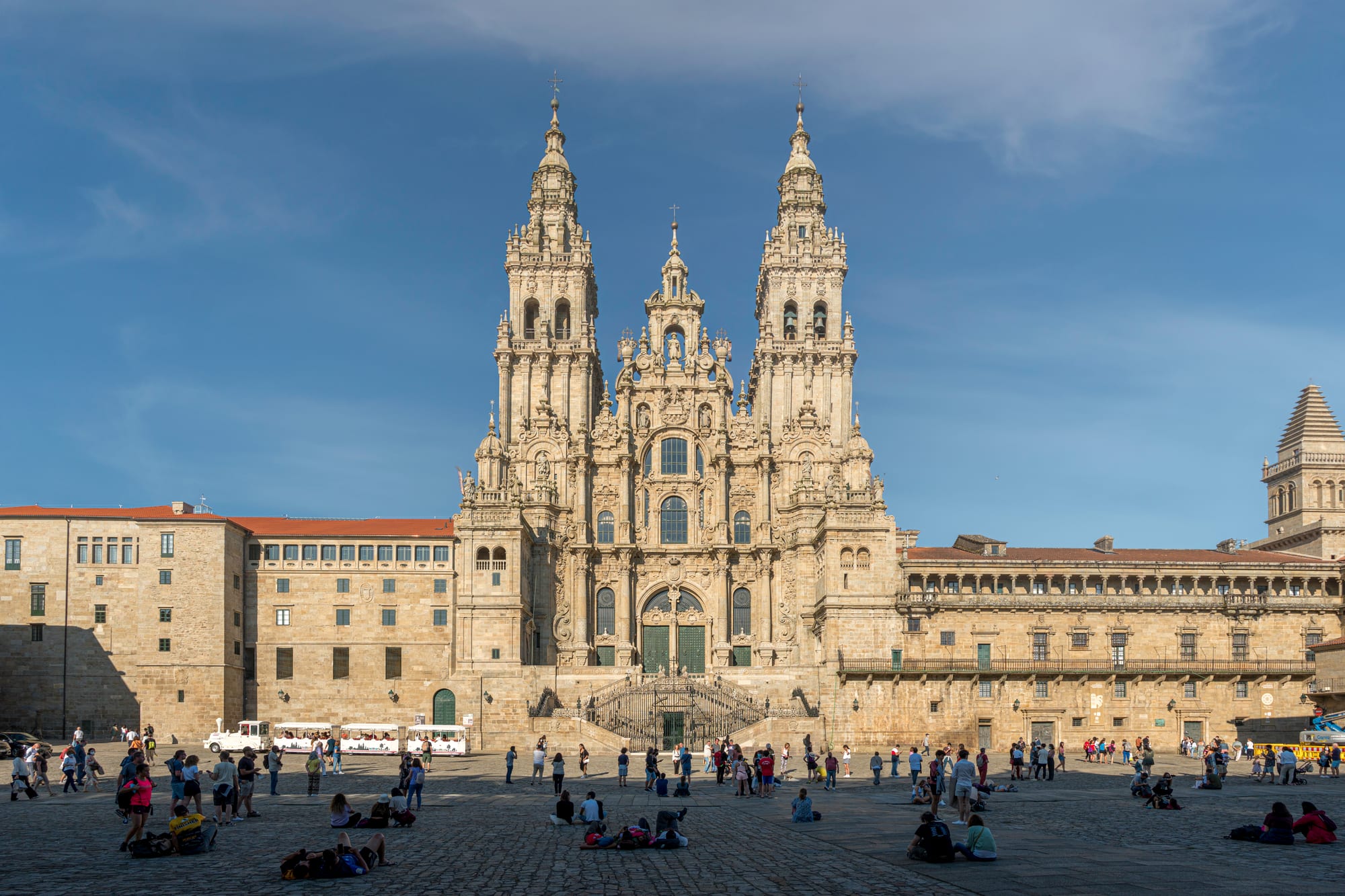 A towering baroque cathedral front with two tall spires above a wide stone square where people sit and rest on the paving