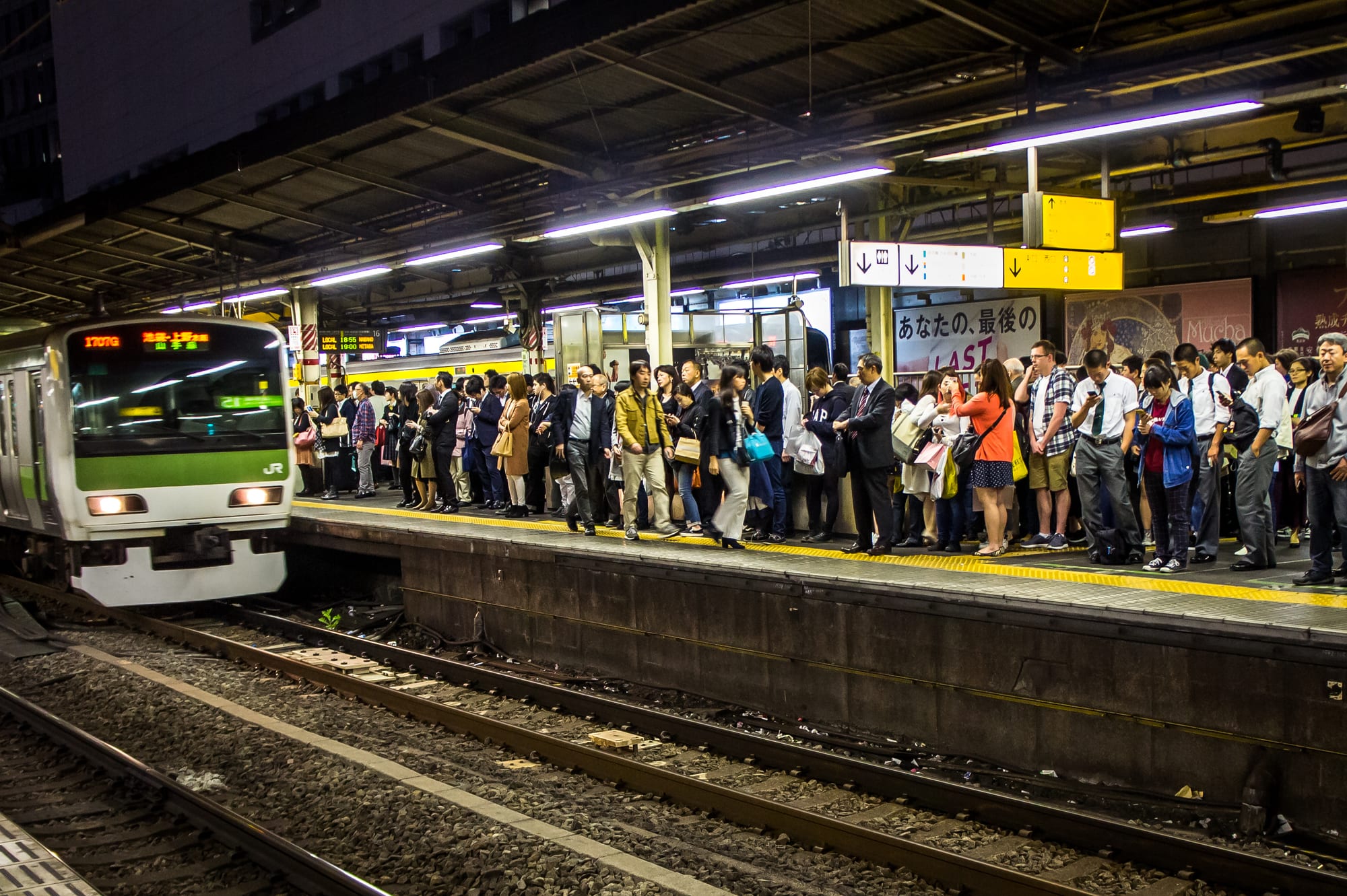 Crowds of commuters in neat lines along a station platform as a green-striped train pulls in under a long roof