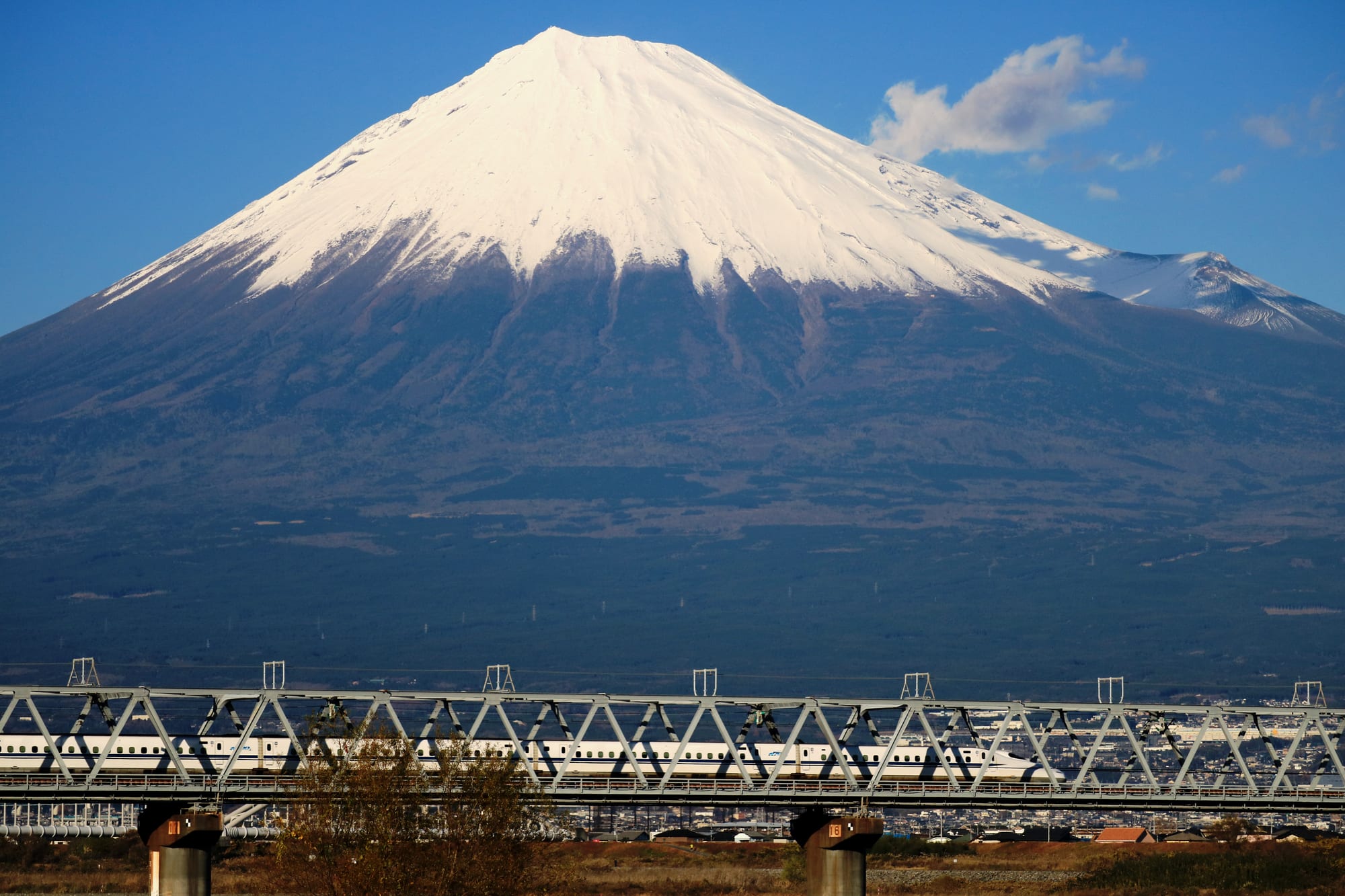 A white high-speed train crossing a long steel truss bridge, with snow-capped Mount Fuji filling the sky behind