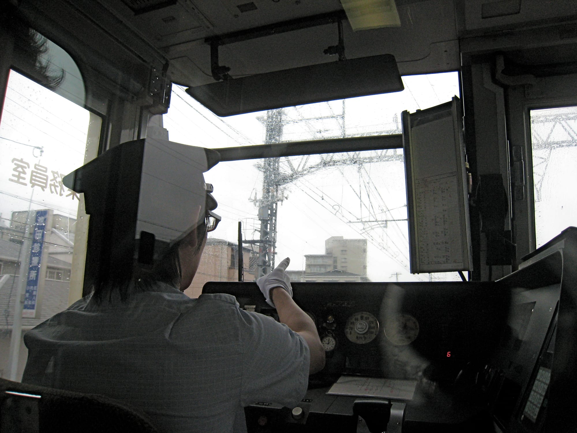 A train driver in a white cap and gloves seated in a cab, pointing ahead through a rain-spotted windscreen at overhead wires and buildings