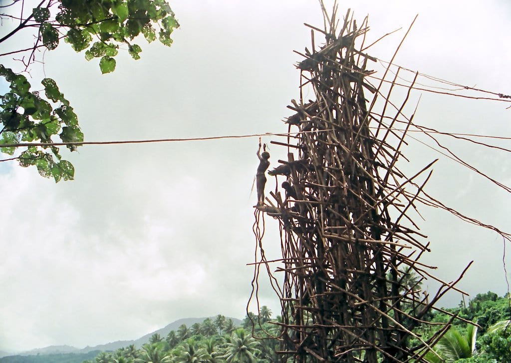 A tall, shaggy tower of lashed wooden poles under a cloudy sky, with a man standing on a platform near the middle and green hills behind