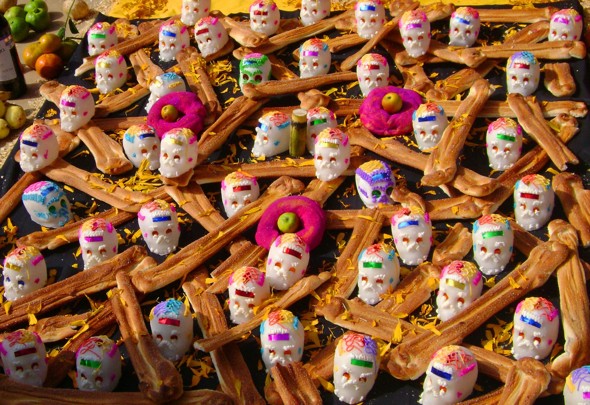 Rows of small white sugar skulls with coloured icing laid out among long bone-shaped loaves of bread on a black cloth scattered with marigold petals