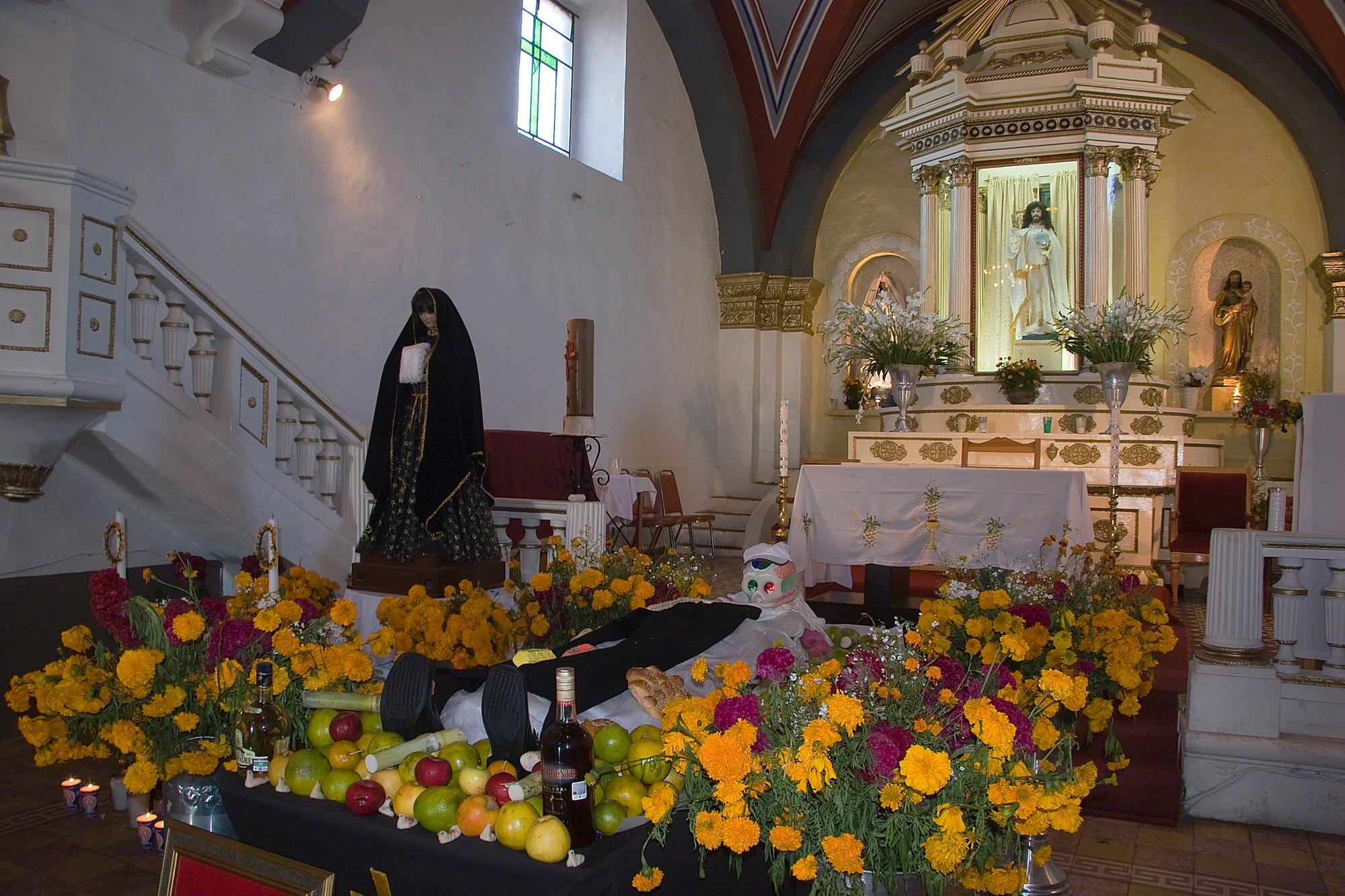 Banks of orange and yellow marigolds, fruit, bread, bottles and candles laid out before the altar of a white church interior