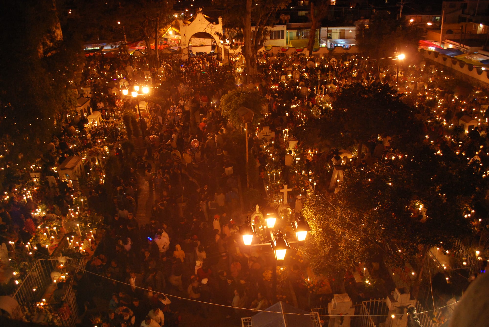 A crowded cemetery at night seen from above, its graves lit by hundreds of candles among trees and lamp posts