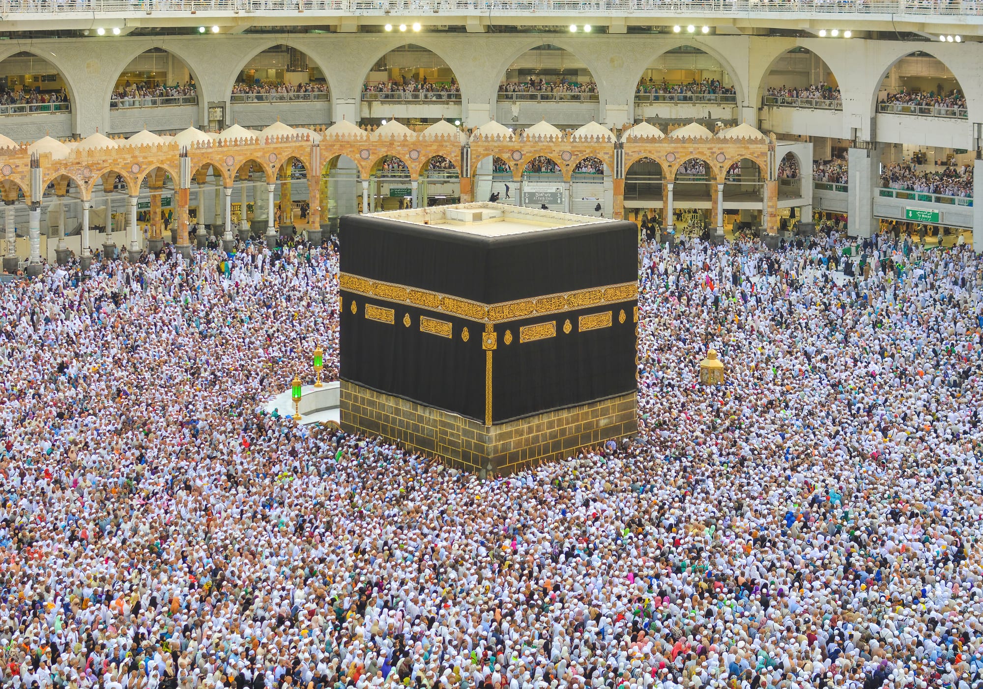A tall black cube draped in cloth with a band of gold embroidery, surrounded on all sides by a dense crowd of pilgrims mostly dressed in white, inside a vast arcaded courtyard