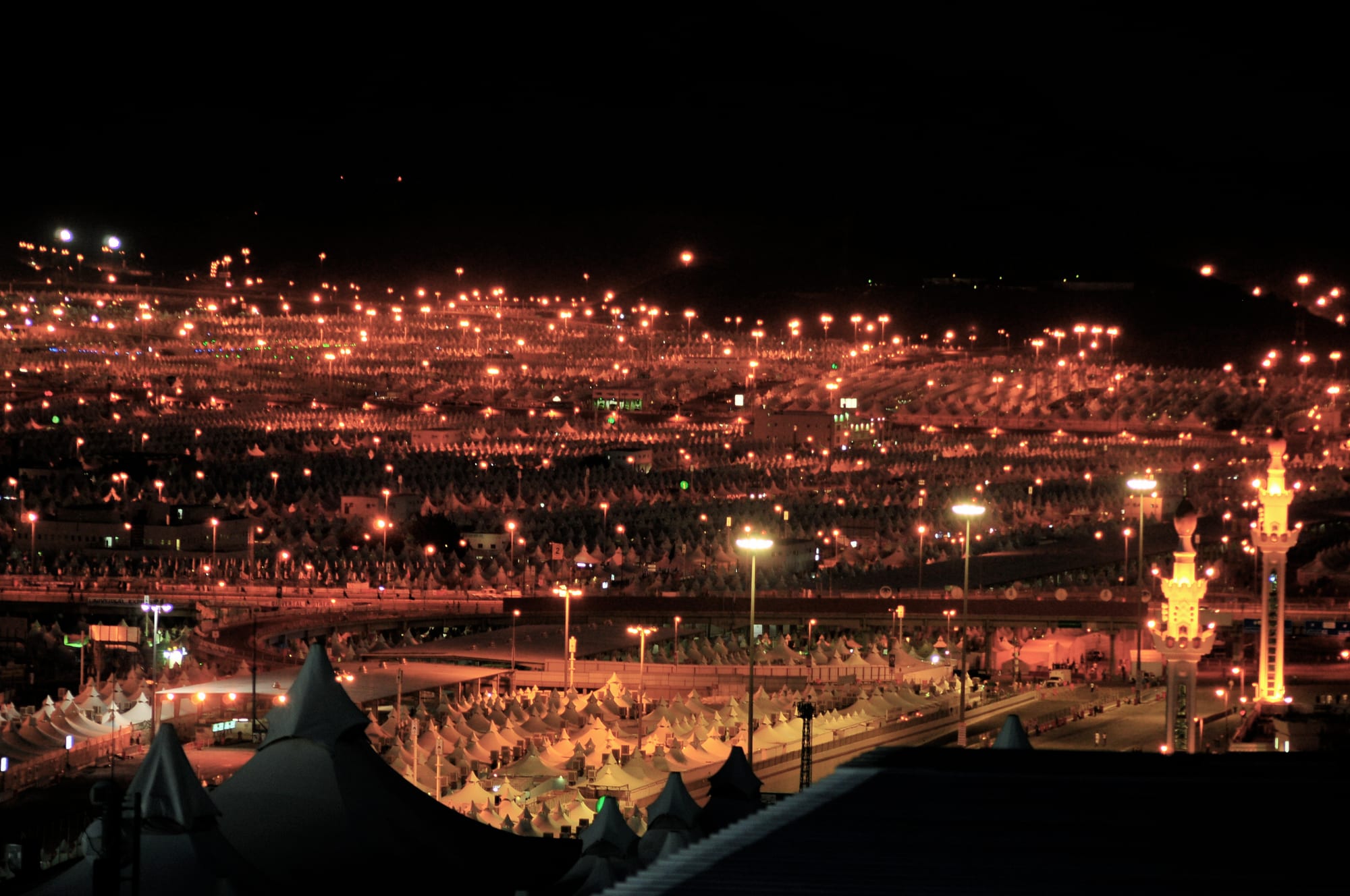 A valley at night filled with thousands of rows of white tents lit by orange street lamps, with a lit minaret on the right