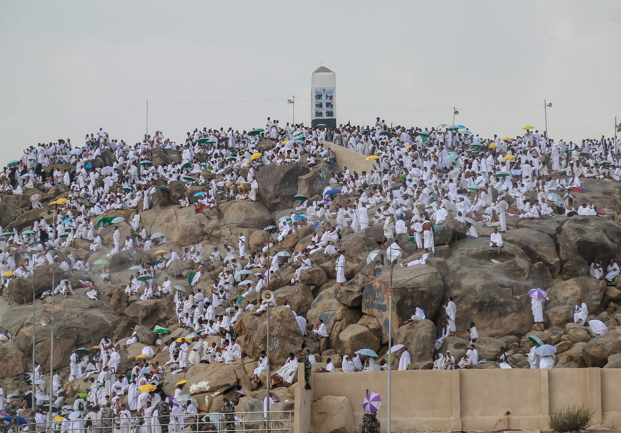 A rocky hill covered with thousands of pilgrims in white robes, many holding coloured umbrellas, with a white pillar on the summit