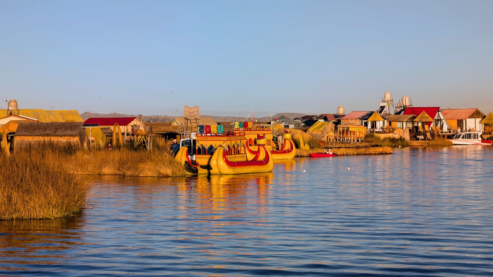 A large yellow and red reed boat moored beside low reed islands with thatched and red-roofed houses, reflected in calm water in evening light