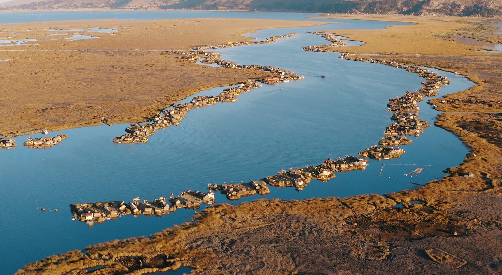 An aerial view of a winding blue channel lined with dozens of small reed islands and huts, surrounded by golden reed beds