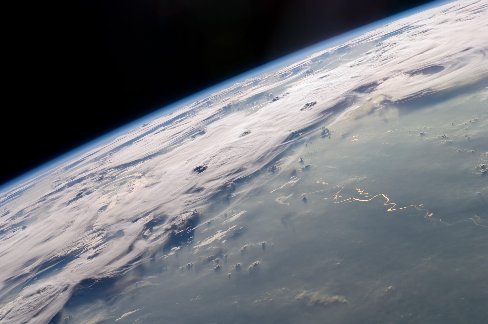 Lines of white storm clouds over a green landscape seen from orbit, with a winding river glinting below