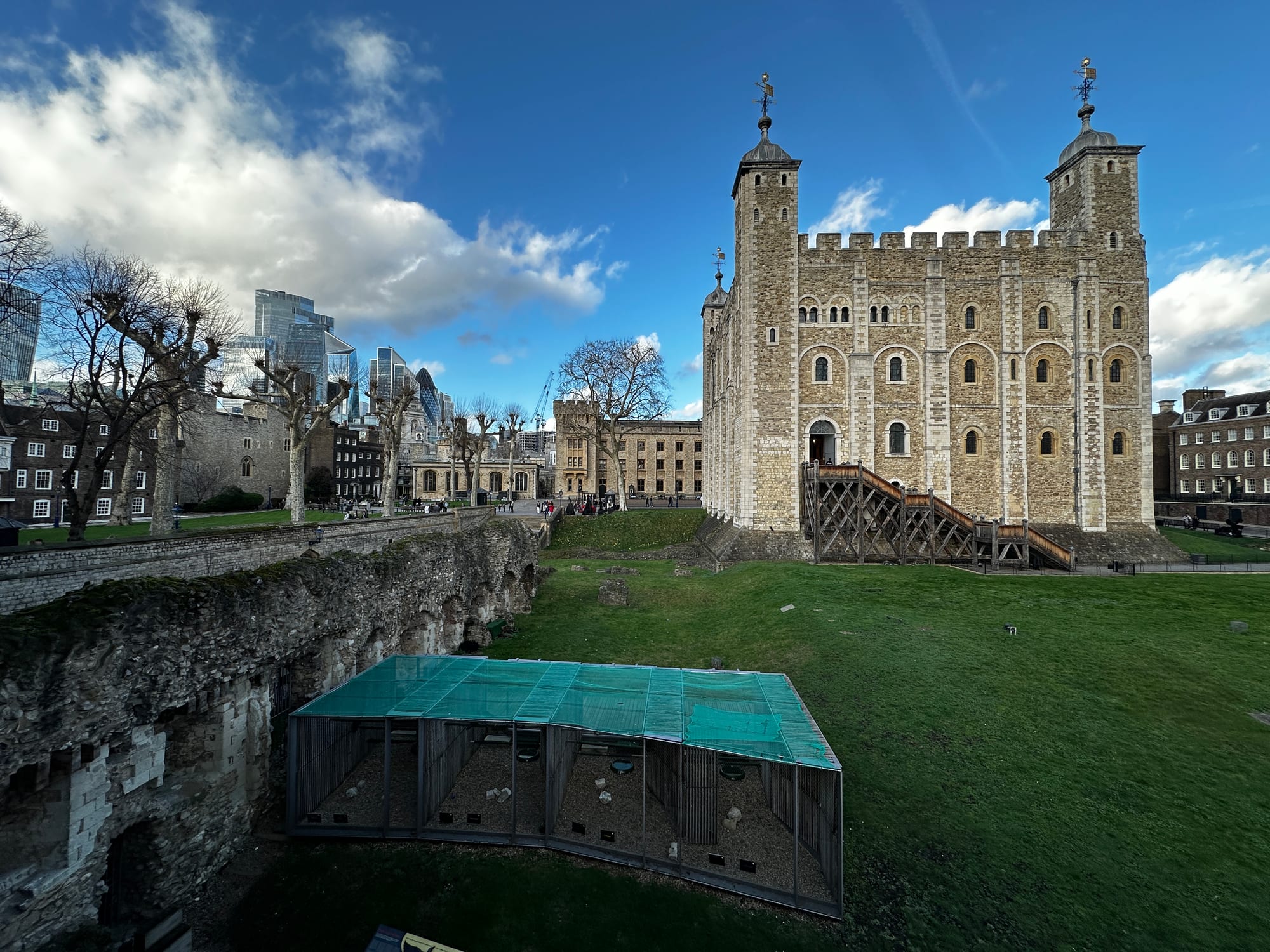A low enclosure with a green mesh roof sits on a lawn beside old stone walls, with a tall square stone keep and a wooden staircase behind it