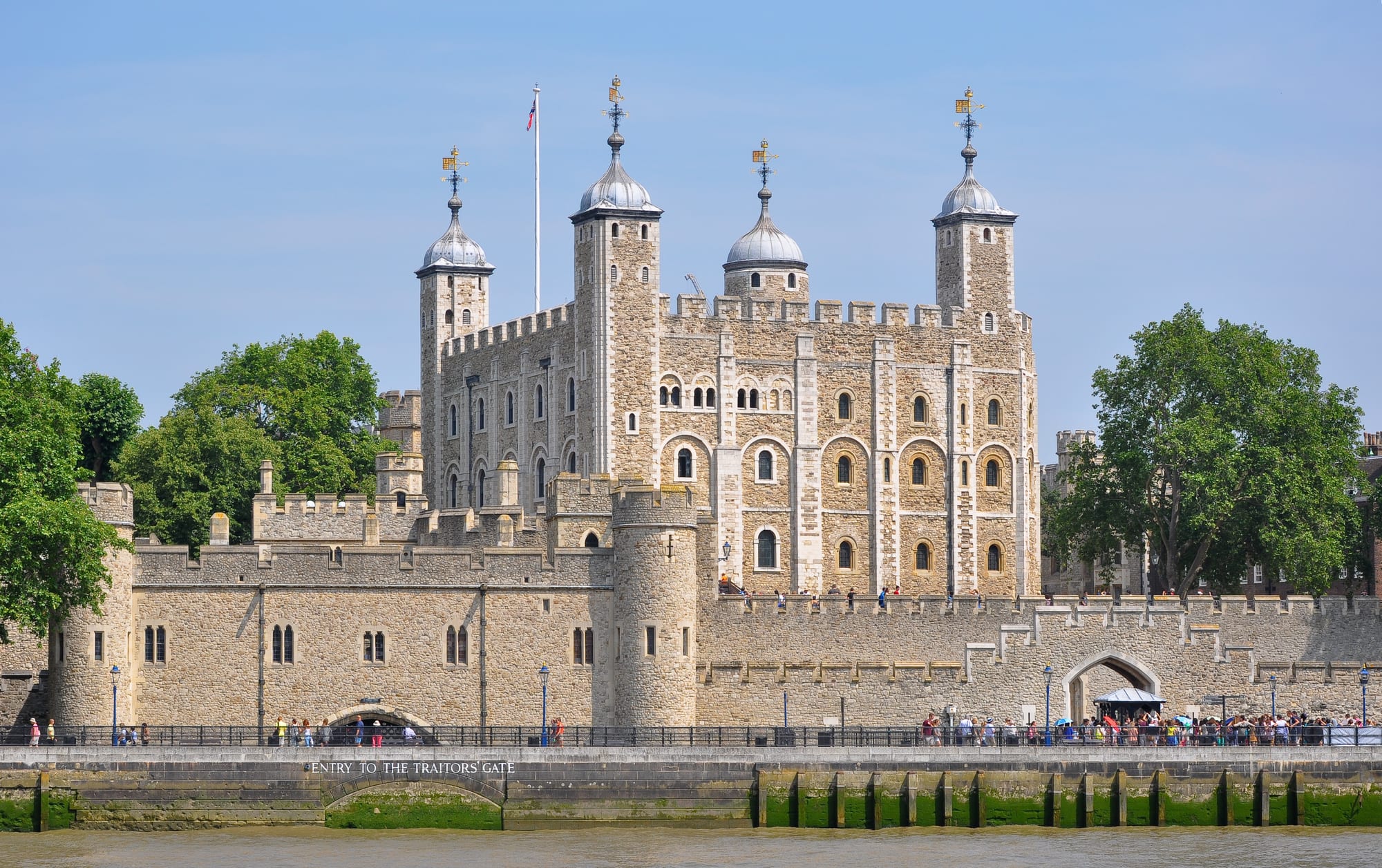 A square, pale stone keep with four corner turrets topped by lead domes and weather vanes rises behind battlemented outer walls on the bank of a river
