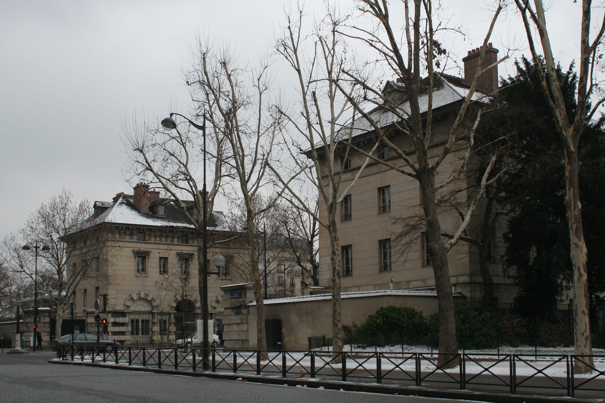 Two square stone pavilions with snowy roofs behind bare plane trees and iron railings on a city square