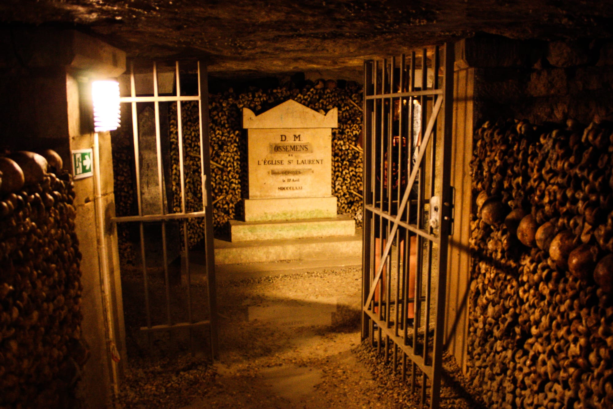 An underground passage lined on both sides with walls of stacked skulls and bones, leading through an iron gate to a carved stone tablet