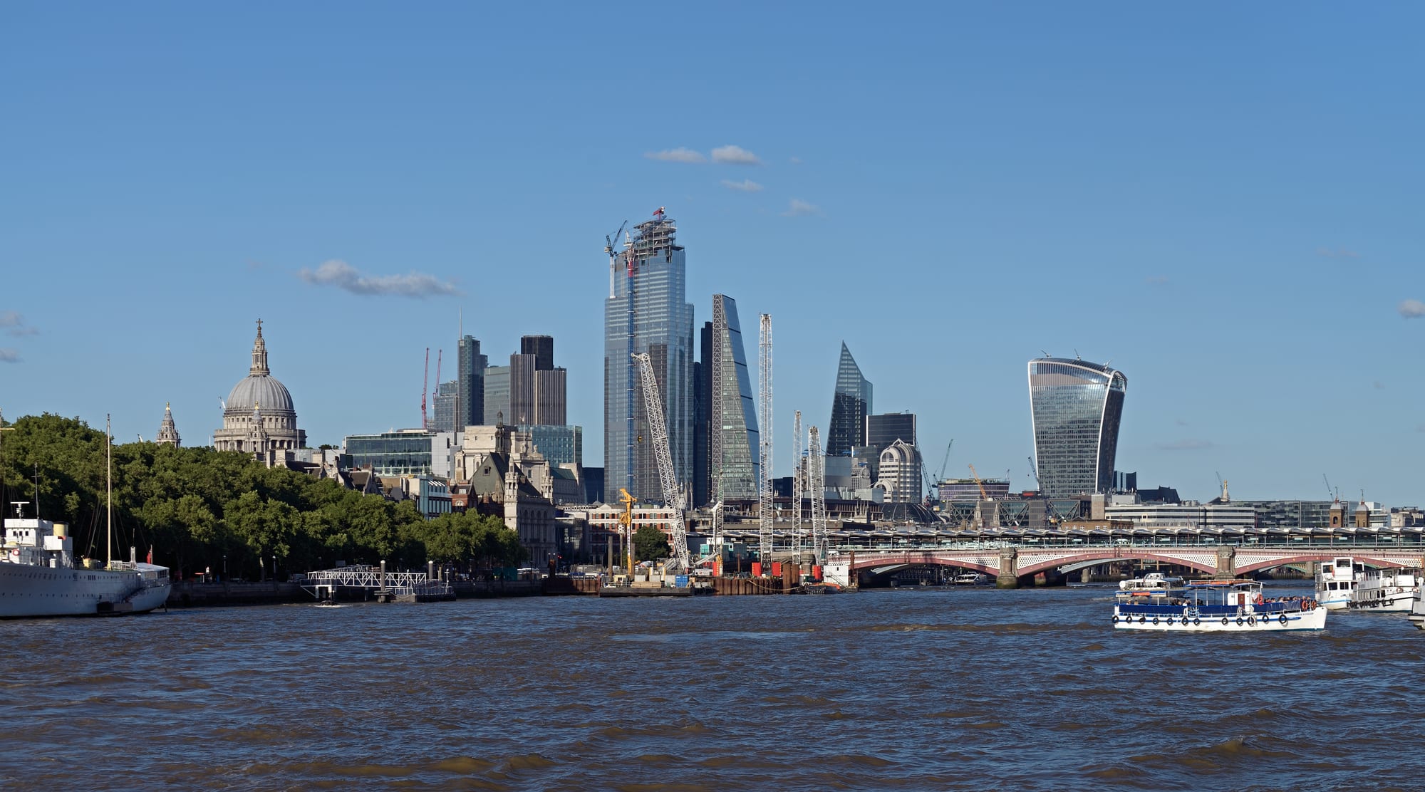 A river in the foreground with a domed cathedral, glass skyscrapers and an arched red bridge behind
