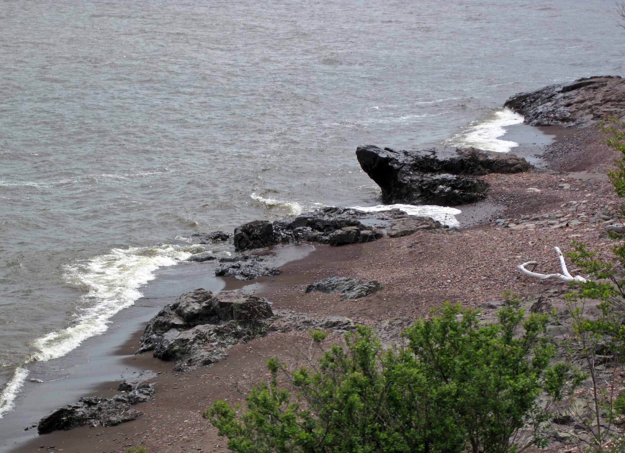 Dark, rounded rocks on a reddish pebble beach where small waves break, with green shrubs in the foreground