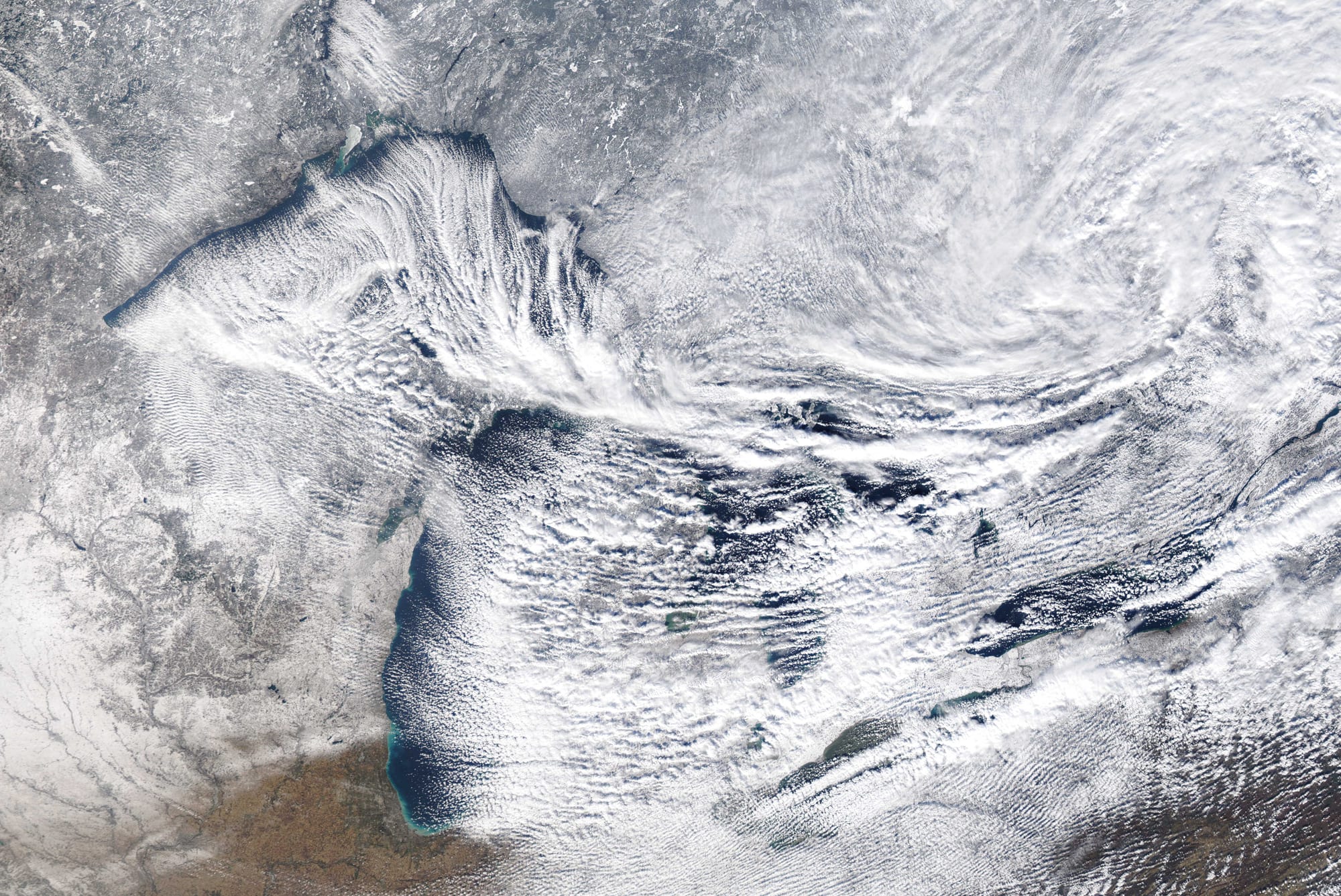 A satellite view of snow-covered land around several large lakes, with long streaks of cloud forming over the open water