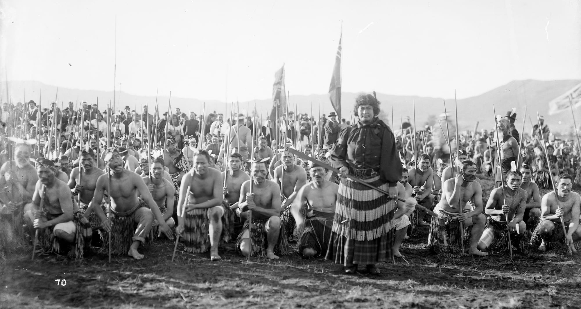 A black-and-white photograph of rows of Māori men kneeling with long staffs, a woman standing in a striped cloak at the centre
