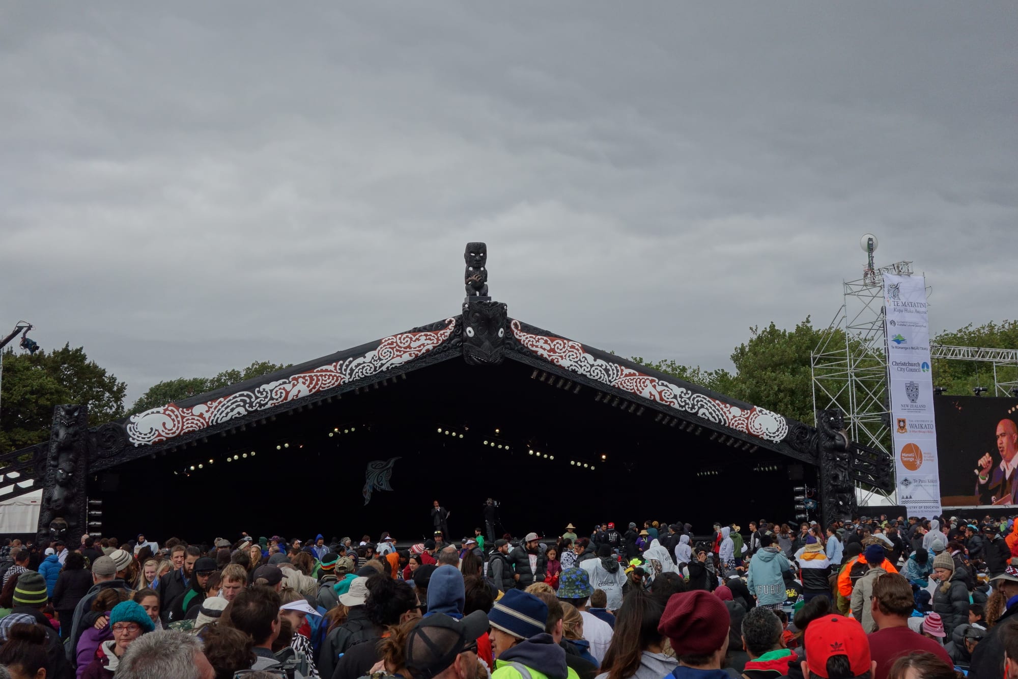 A large crowd in front of an outdoor stage whose roof is carved and painted in red, white and black Māori patterns