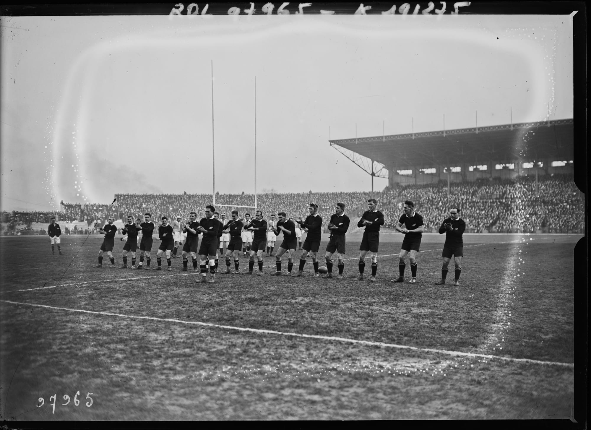 A black-and-white photograph of a line of rugby players in dark kit performing a haka in front of a packed grandstand