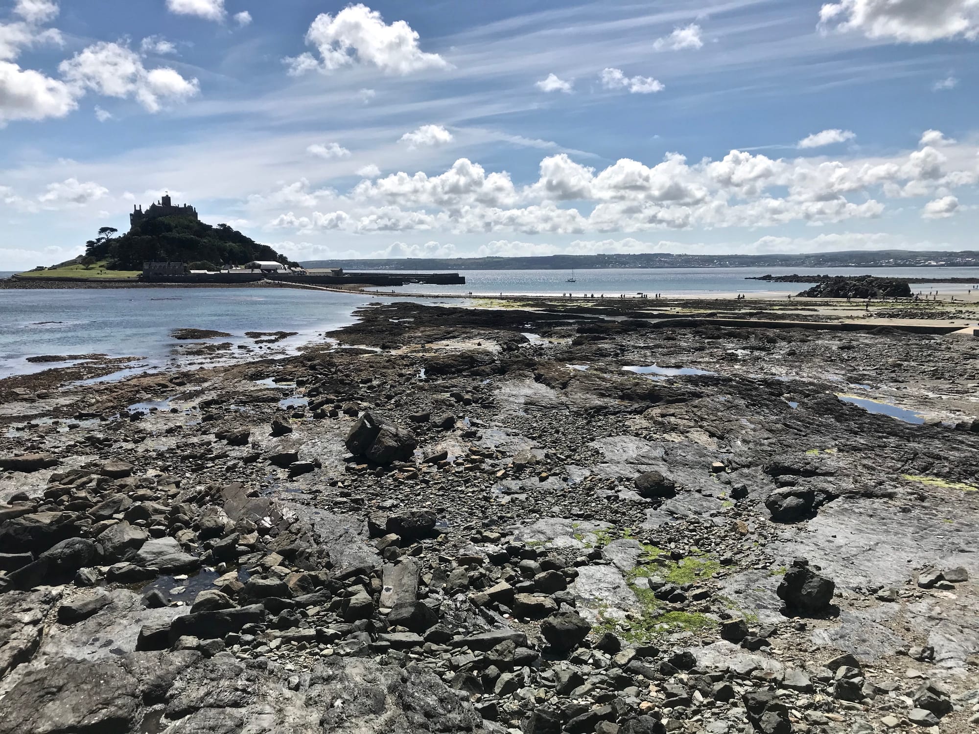 A castle-topped island beyond a wide stretch of exposed rocks and a stone causeway at low tide, under a blue sky with white clouds